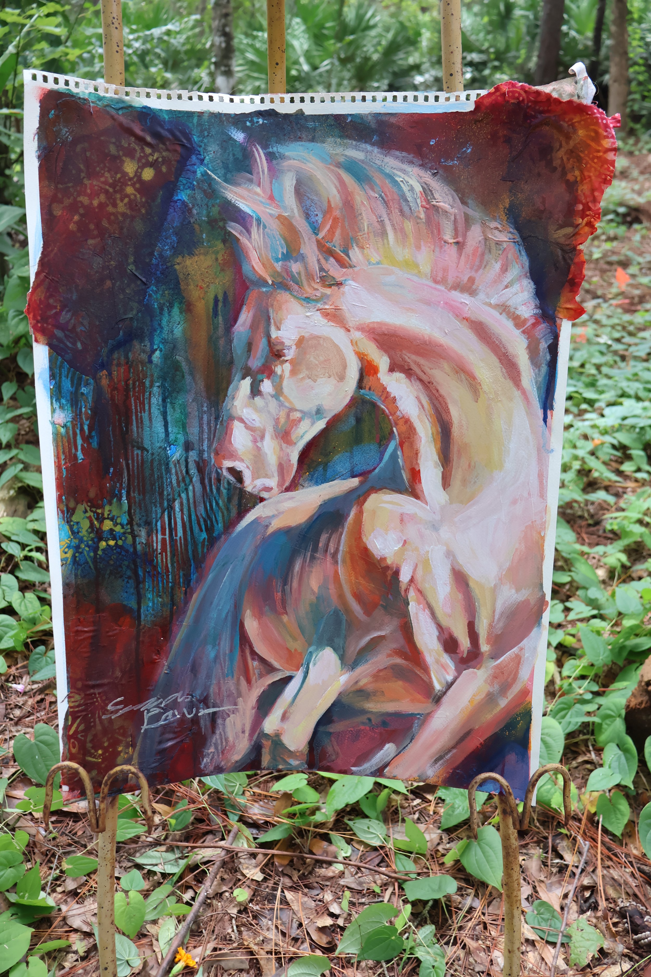 Colorful abstract painting of a seated figure displayed outdoors among trees and forest vegetation