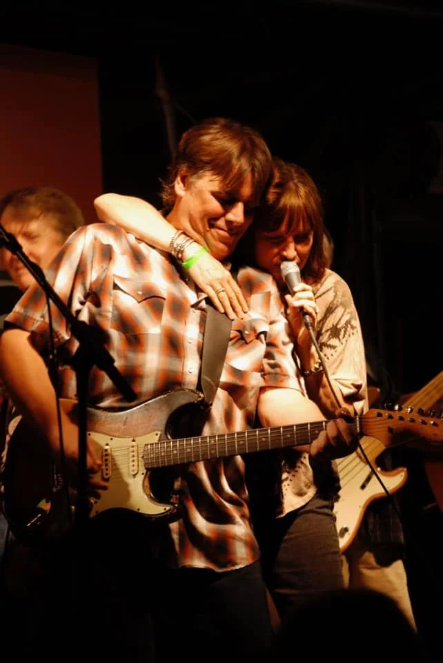 Two musicians, one playing an electric guitar and the other holding a microphone, embrace on stage during a live performance with warm lighting.