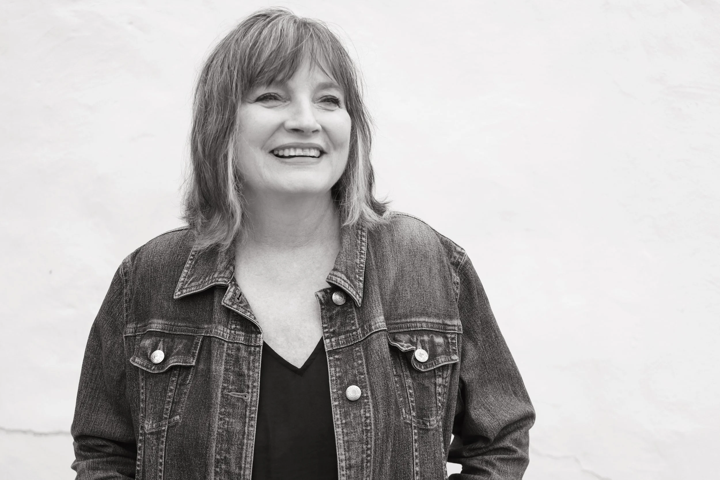 A black and white portrait of a smiling woman with shoulder-length hair, wearing a denim jacket over a dark top, standing against a plain light-colored wall.