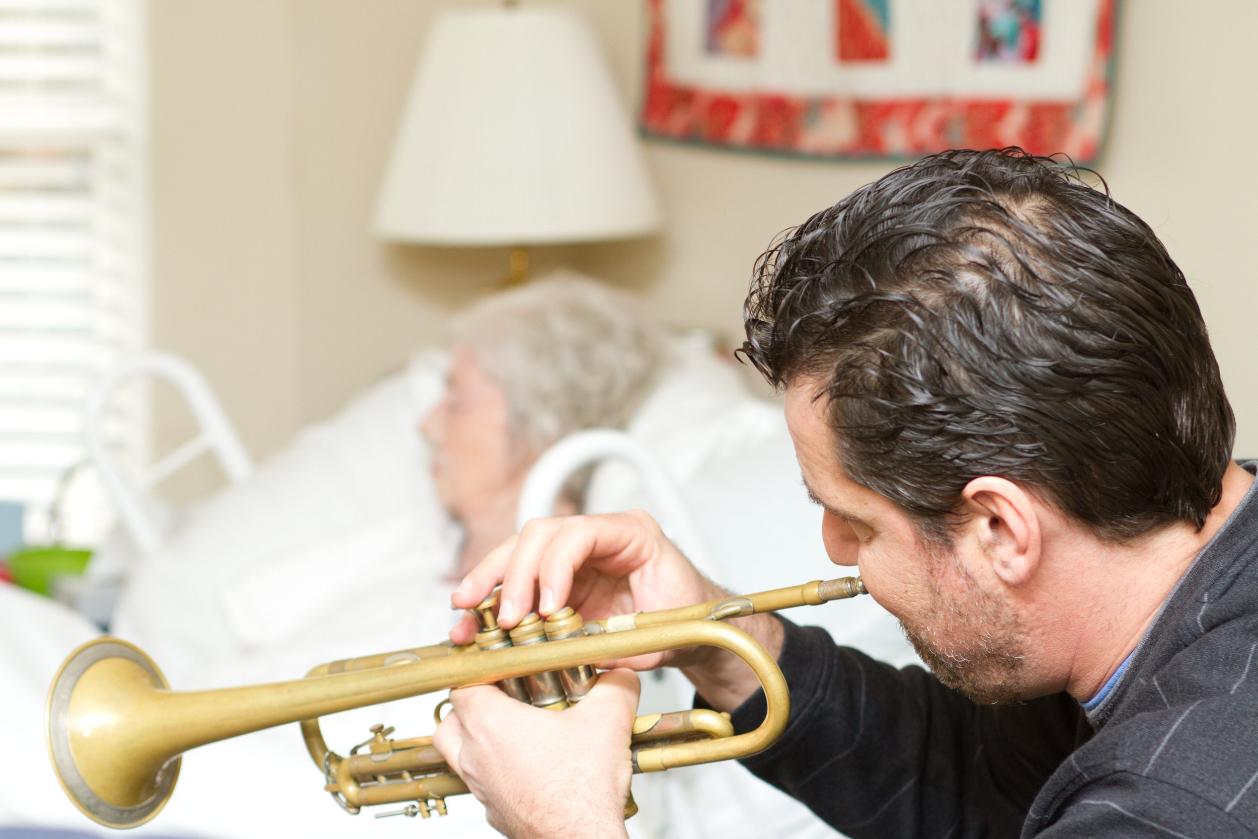 A man playing a trumpet indoors with a woman in bed in the background.