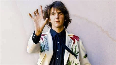 A young man with shoulder-length dark hair making a gesture with his hand, wearing a colorful patterned jacket over a dark shirt, standing against a plain background.