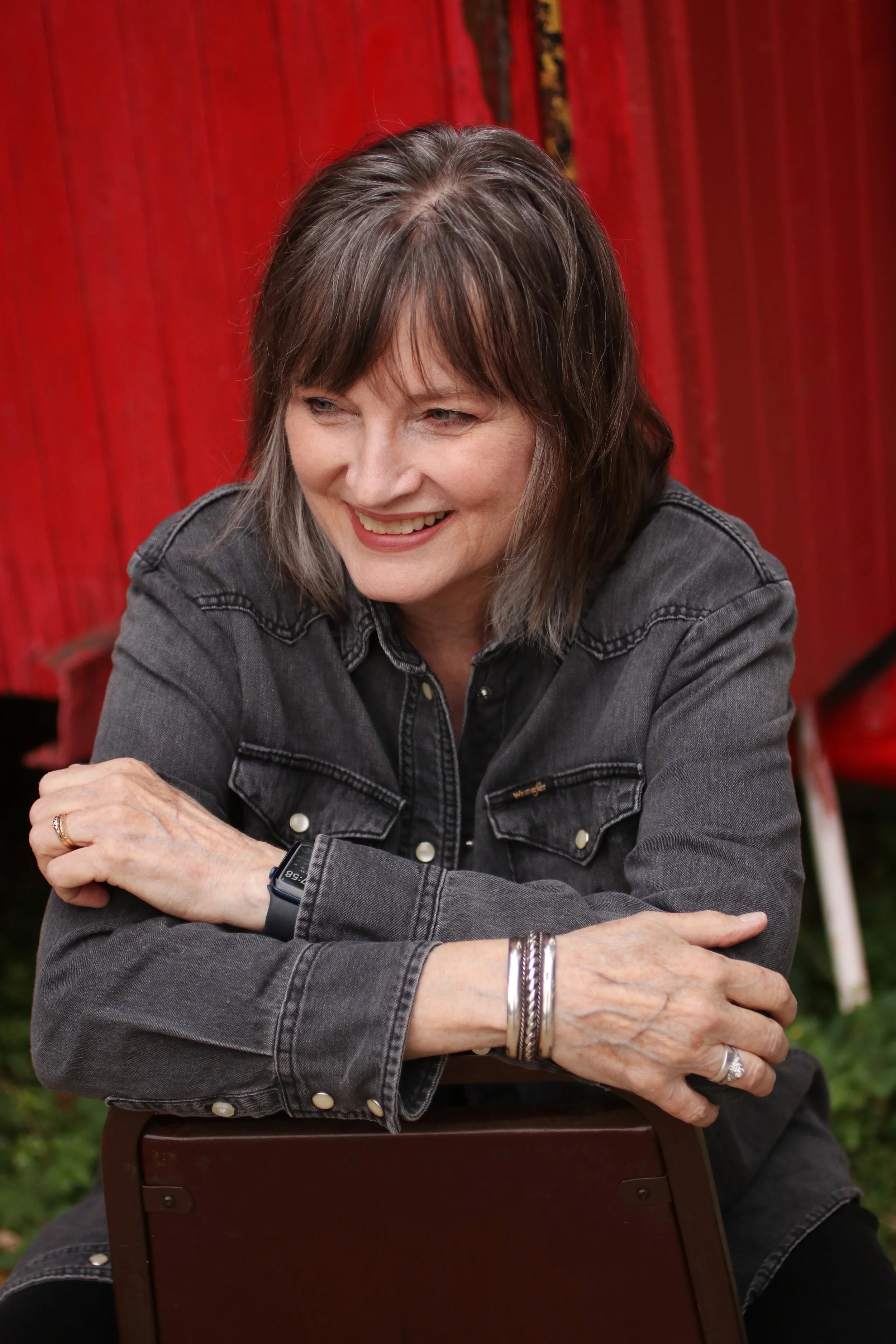 A middle-aged woman with shoulder-length brown hair and bangs smiling. She is wearing a dark denim jacket, a smartwatch on her left wrist, and jewelry on her right wrist. She is seated outdoors against a red wooden background, with her arms crossed resting on the back of a chair.