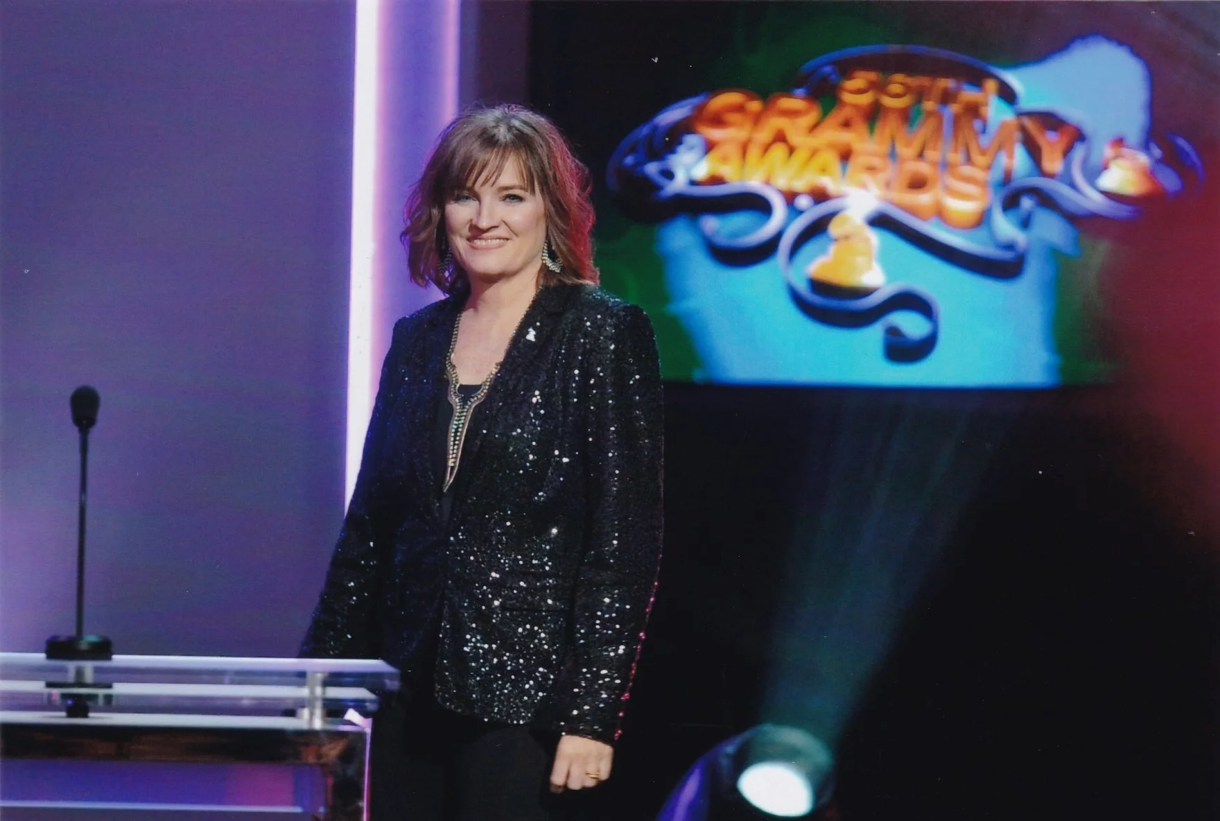 A woman standing on stage wearing a black, sparkly blazer, smiling at the camera. Behind her is a screen with the GRAMMY Awards logo and a microphone on a stand is visible on the left.