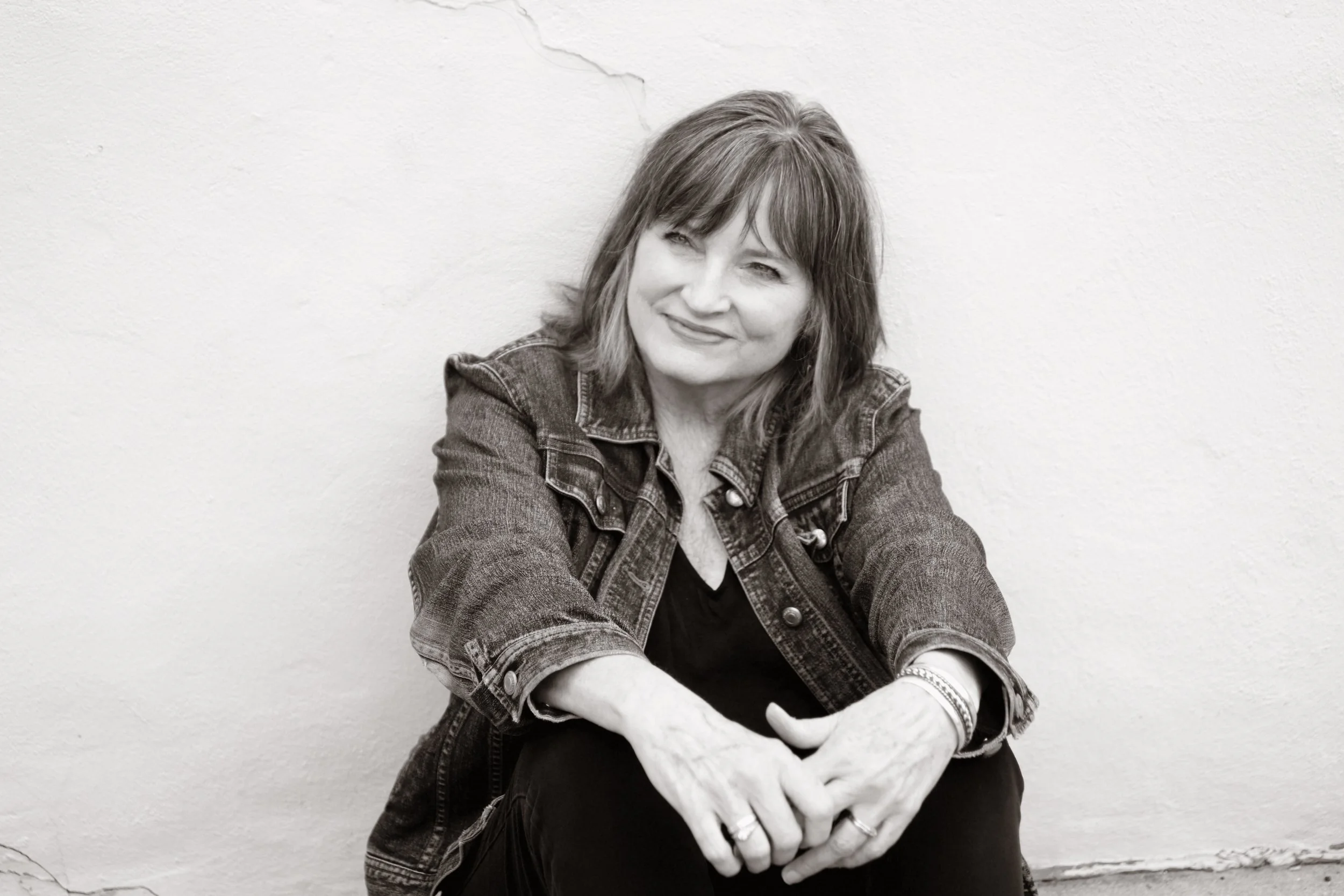 A woman with shoulder-length hair and a denim jacket sitting against a plain wall.
