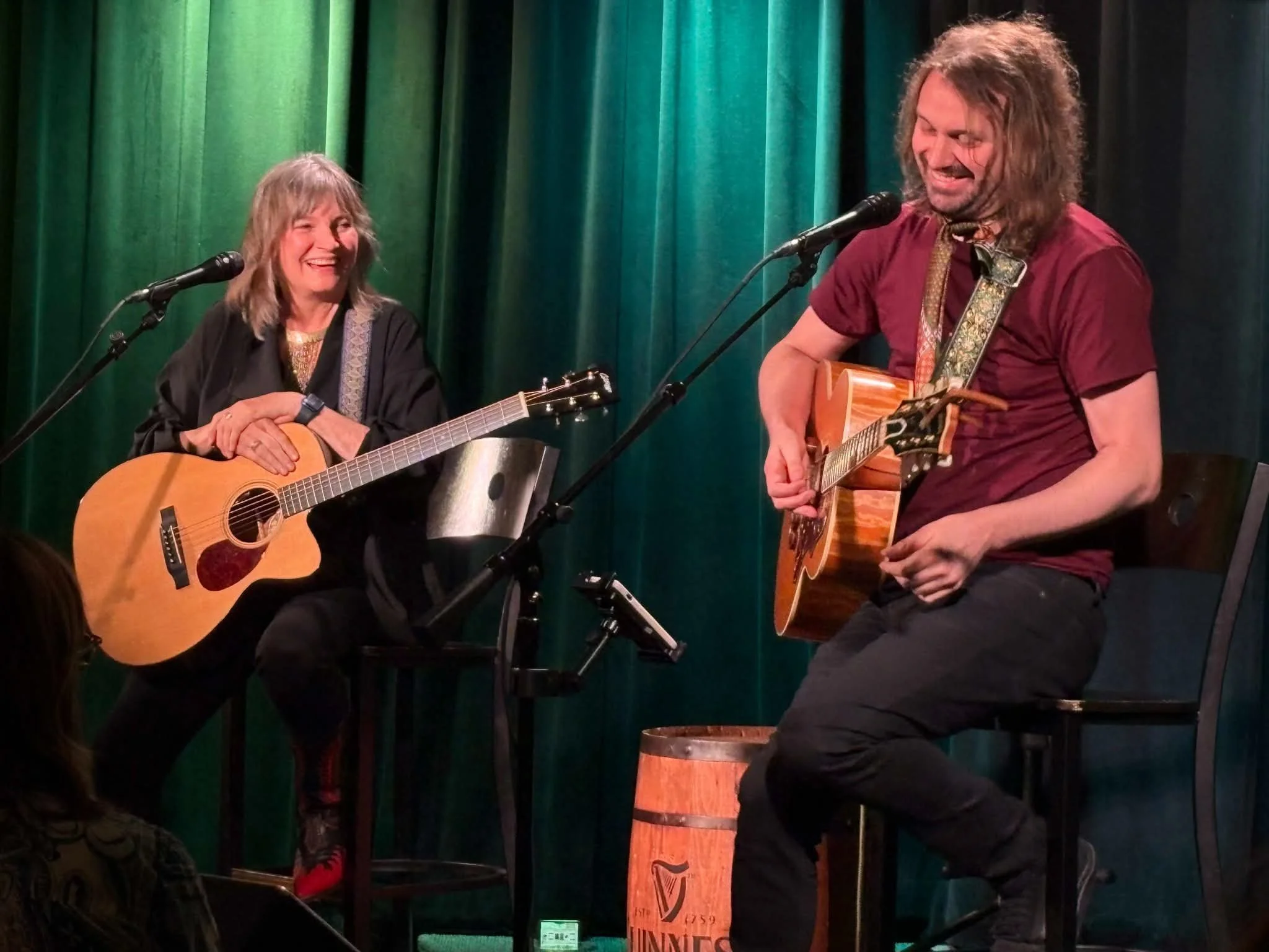 Two musicians performing with acoustic guitars on stage, smiling and enjoying the moment, with a green curtain in the background.