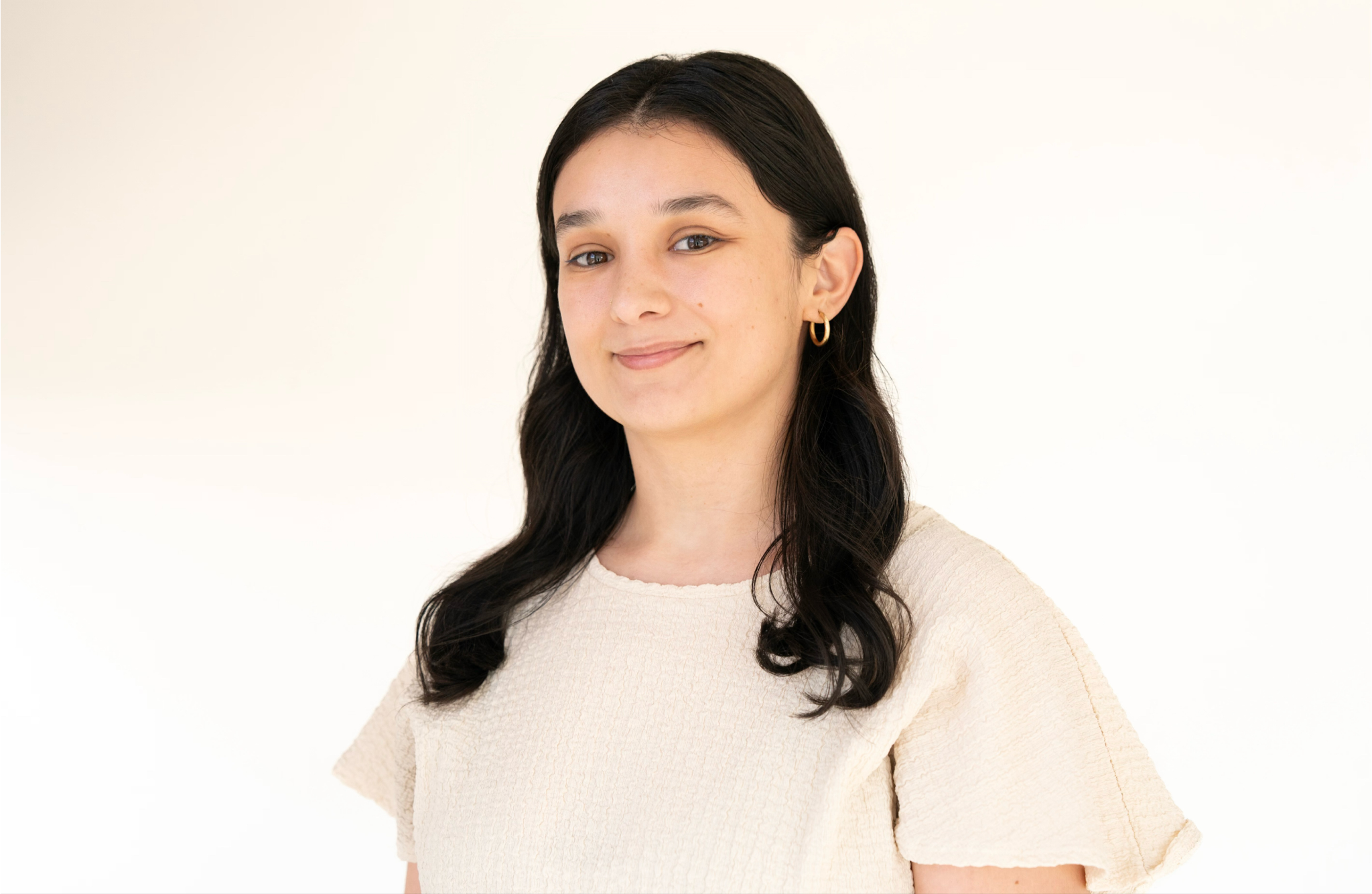 Portrait of a young woman with long dark hair, wearing a cream-colored textured top, against a plain off-white background.