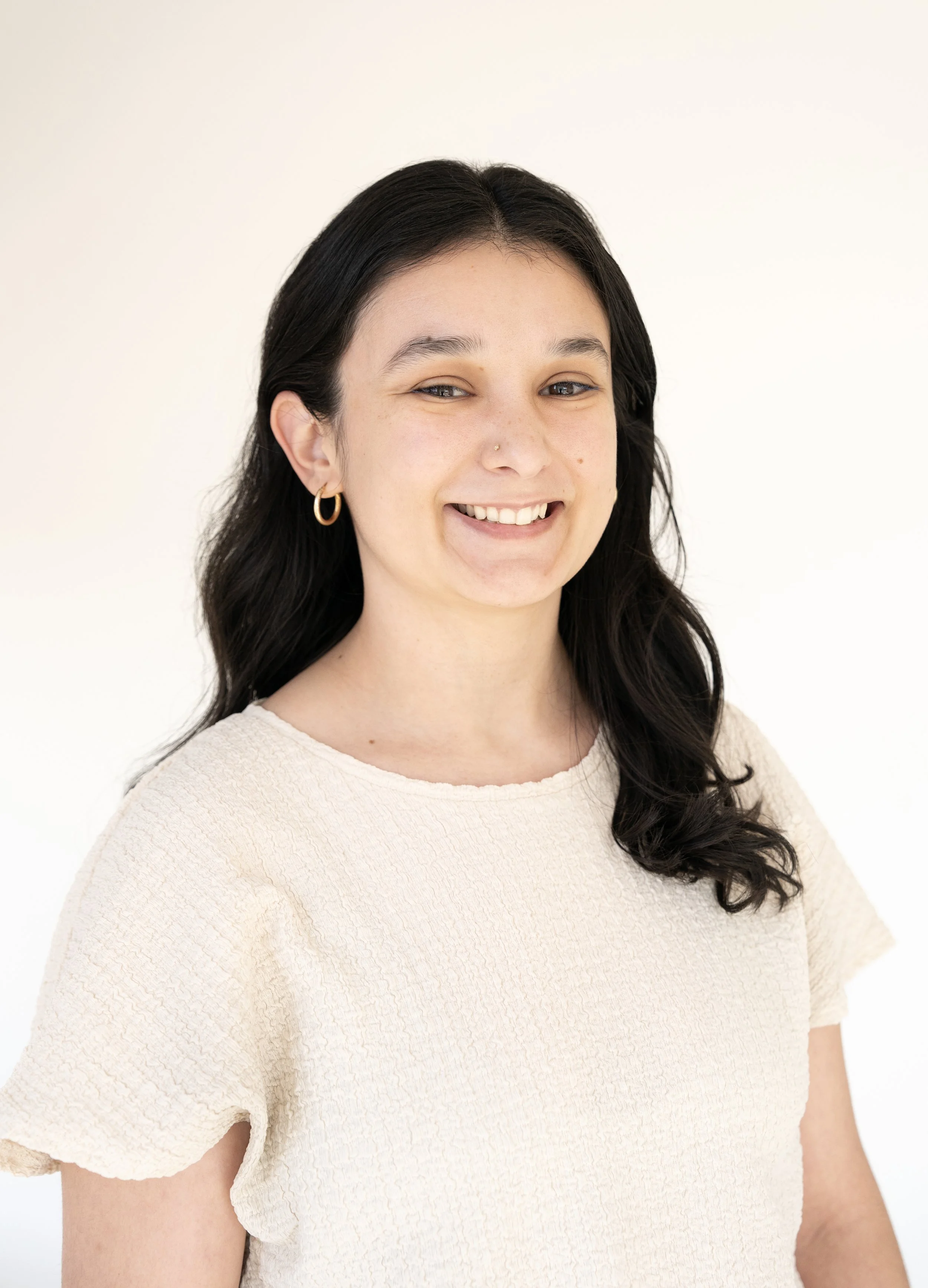 Close-up of a smiling woman with long dark hair, wearing gold hoop earrings and a cream textured top, against a plain white background.