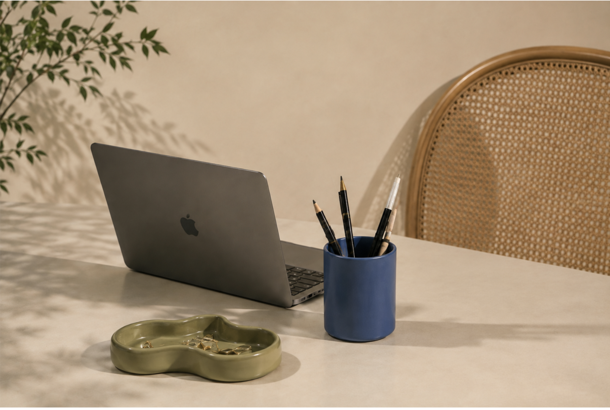 A laptop, a blue container with pens and pencils, a green ceramic dish with jewelry, and a rattan-backed chair in a cozy workspace.