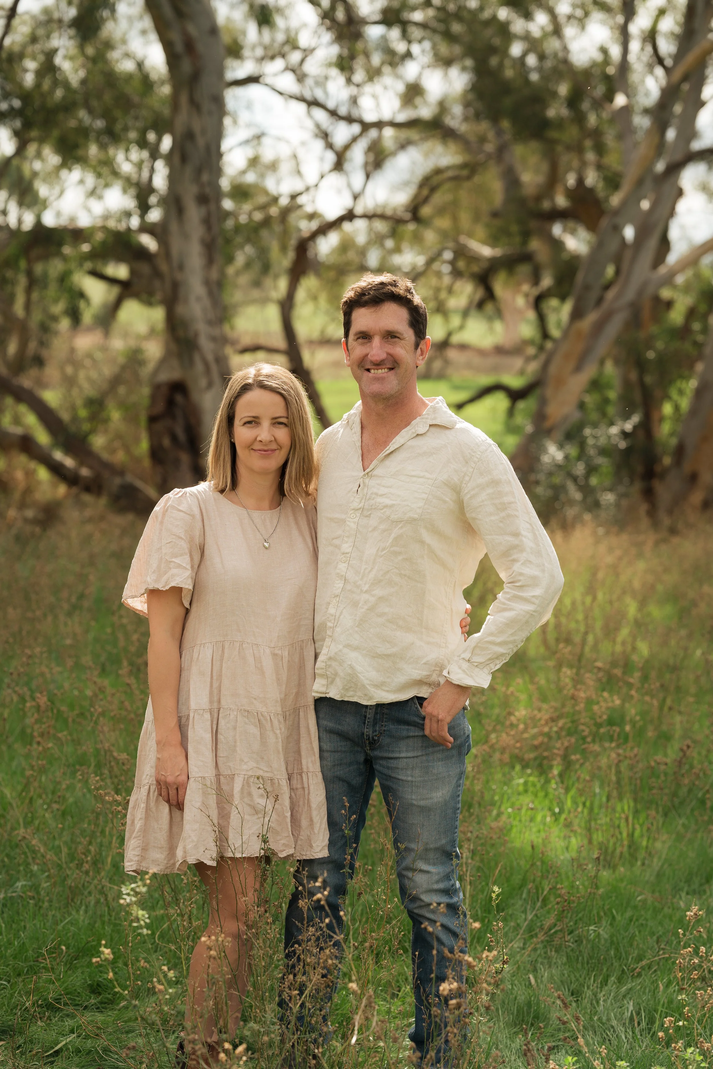 A man and woman standing together outdoors in a grassy area with trees in the background, smiling and looking at the camera.