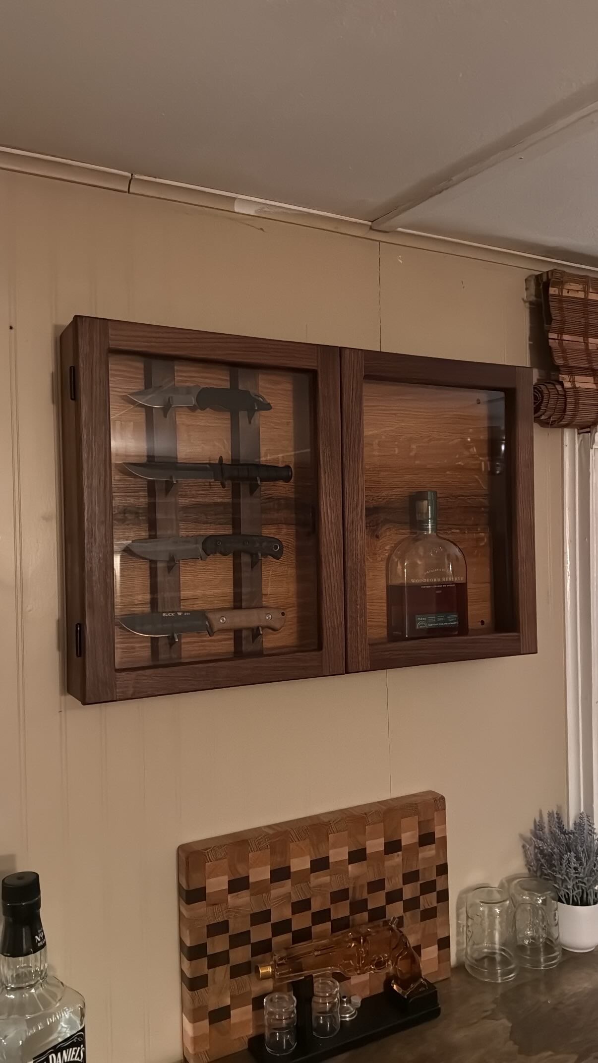 Wooden wall-mounted cabinet with glass doors containing knives and a bottle of whiskey; kitchen countertop below with a glassware set, a whiskey decanter, a cutting board, and a herbal plant in a white pot.