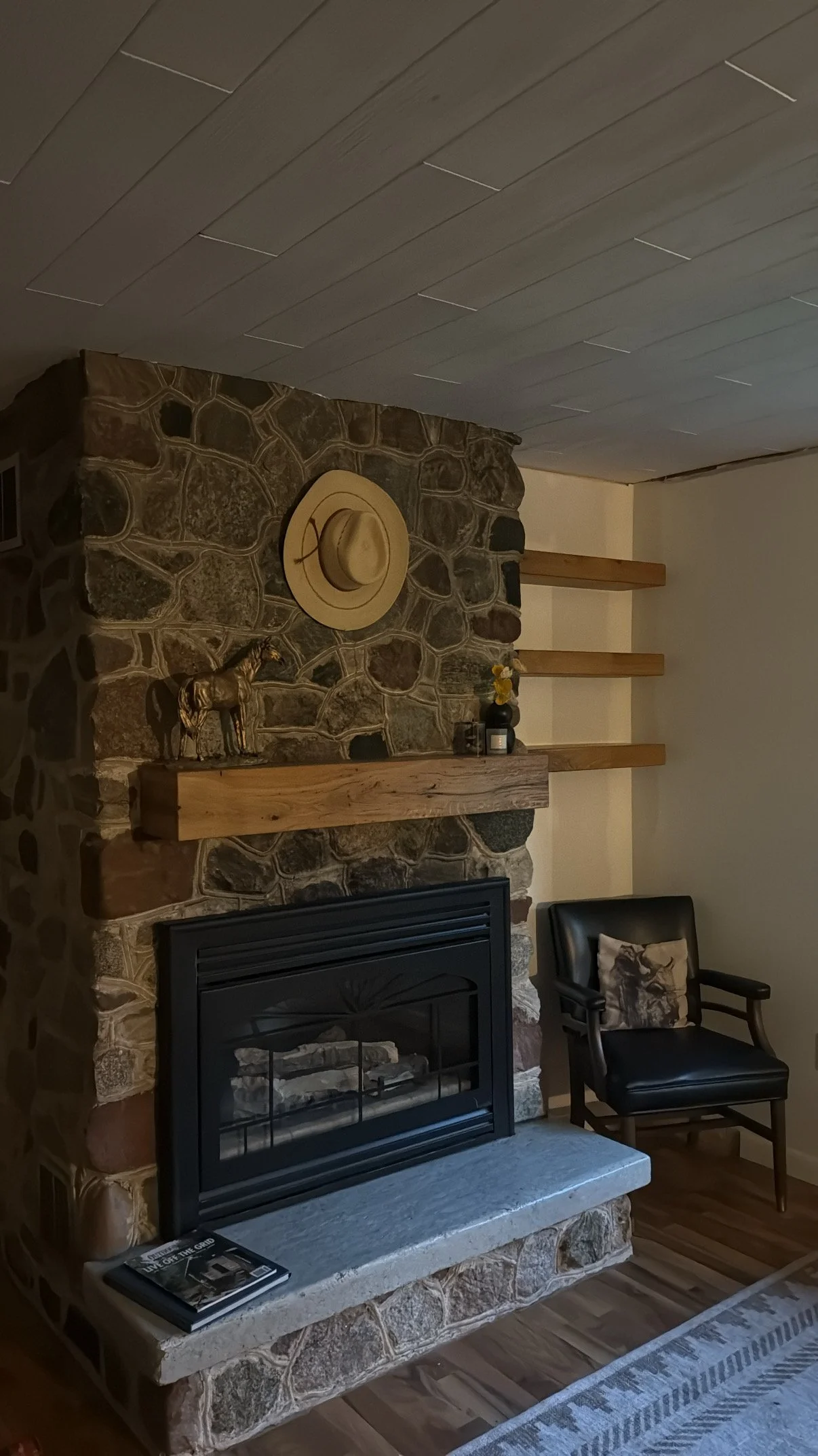 Living room with stone fireplace, wooden mantel, beige wall, black armchair with animal print pillow, and decorative items including a hat, horse figurine, and vase with yellow flowers on mantel.