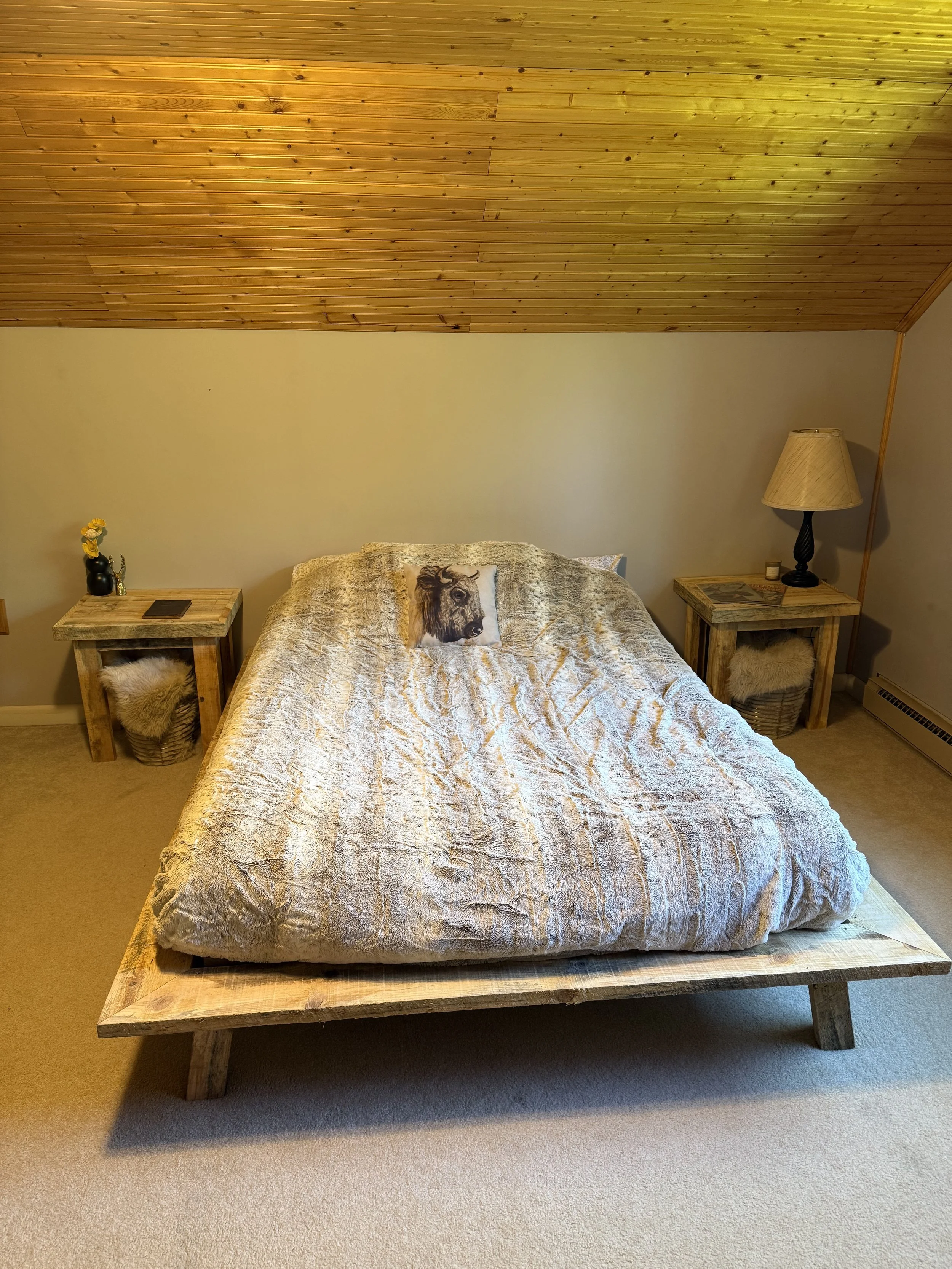 A rustic bedroom with a wooden platform bed, a pillow with a lion's face, two small side tables with lamps, a small vase, and a furry basket, all under a wood-paneled ceiling.