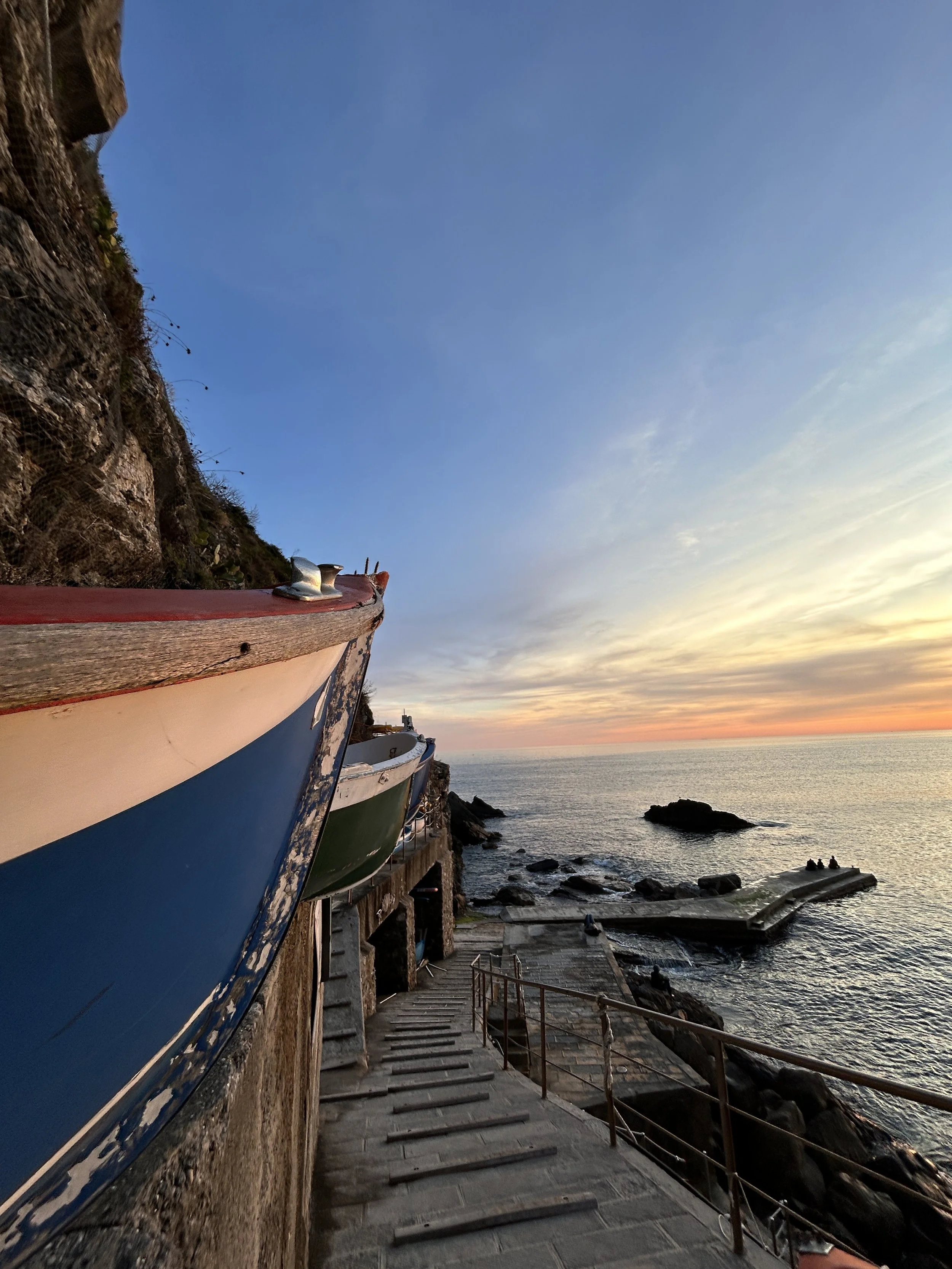 A dockside with colorful boats lined along a stone walkway, overlooking the ocean at sunset, with rocks and waves below.