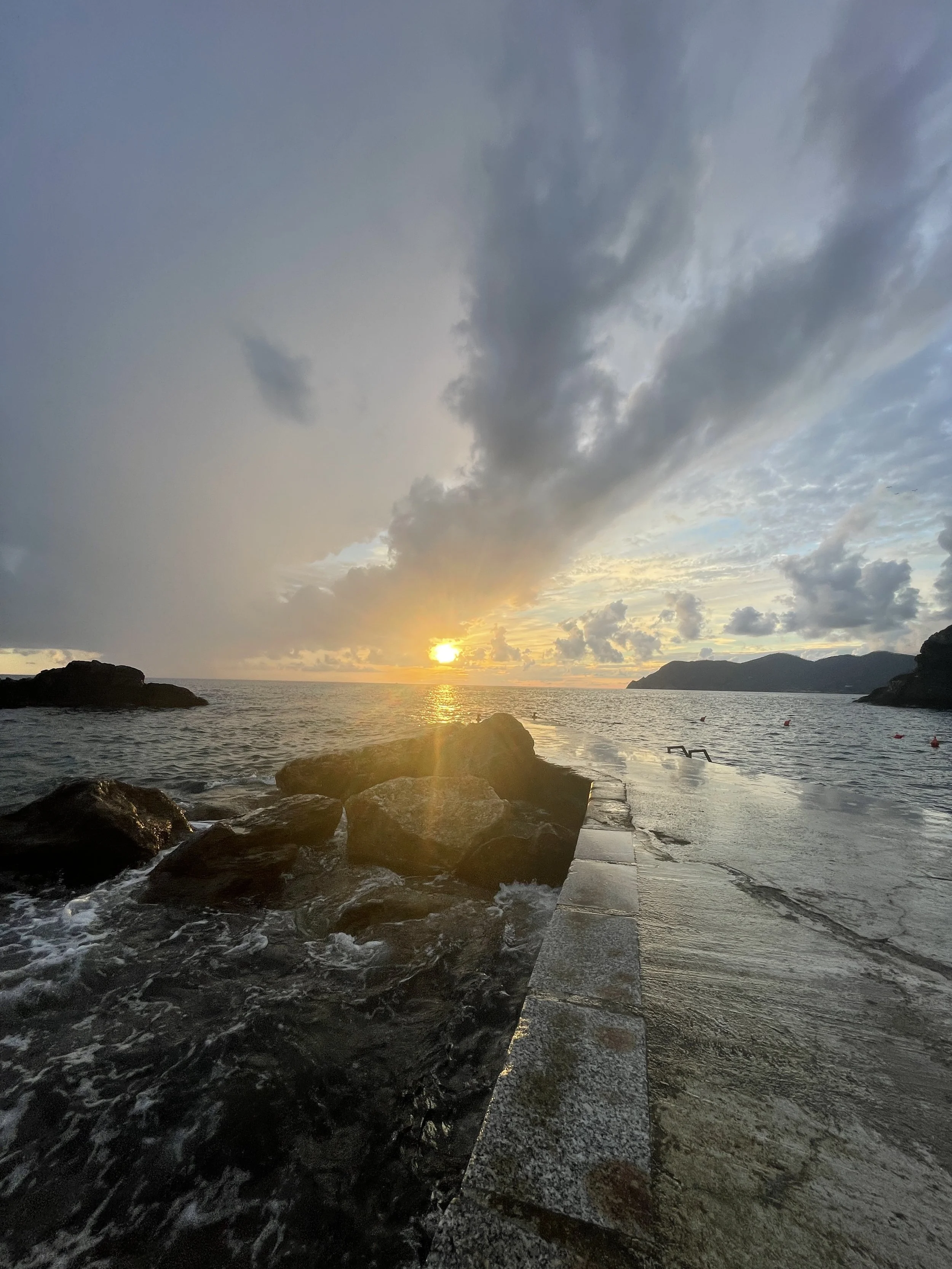 A sunset over the ocean with a rocky shoreline and a stone pier extending into the water. Dark clouds over the magical sky of Corniglia with the sun near the horizon casting a warm glow.