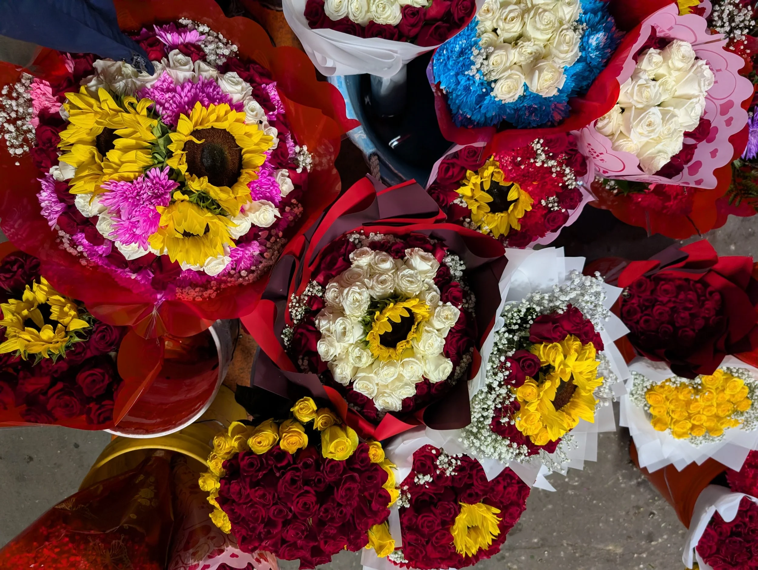 Various bouquets of colorful flowers, including roses, sunflowers, baby’s breath, and chrysanthemums, wrapping in red, white, and pink paper, arranged for sale.
