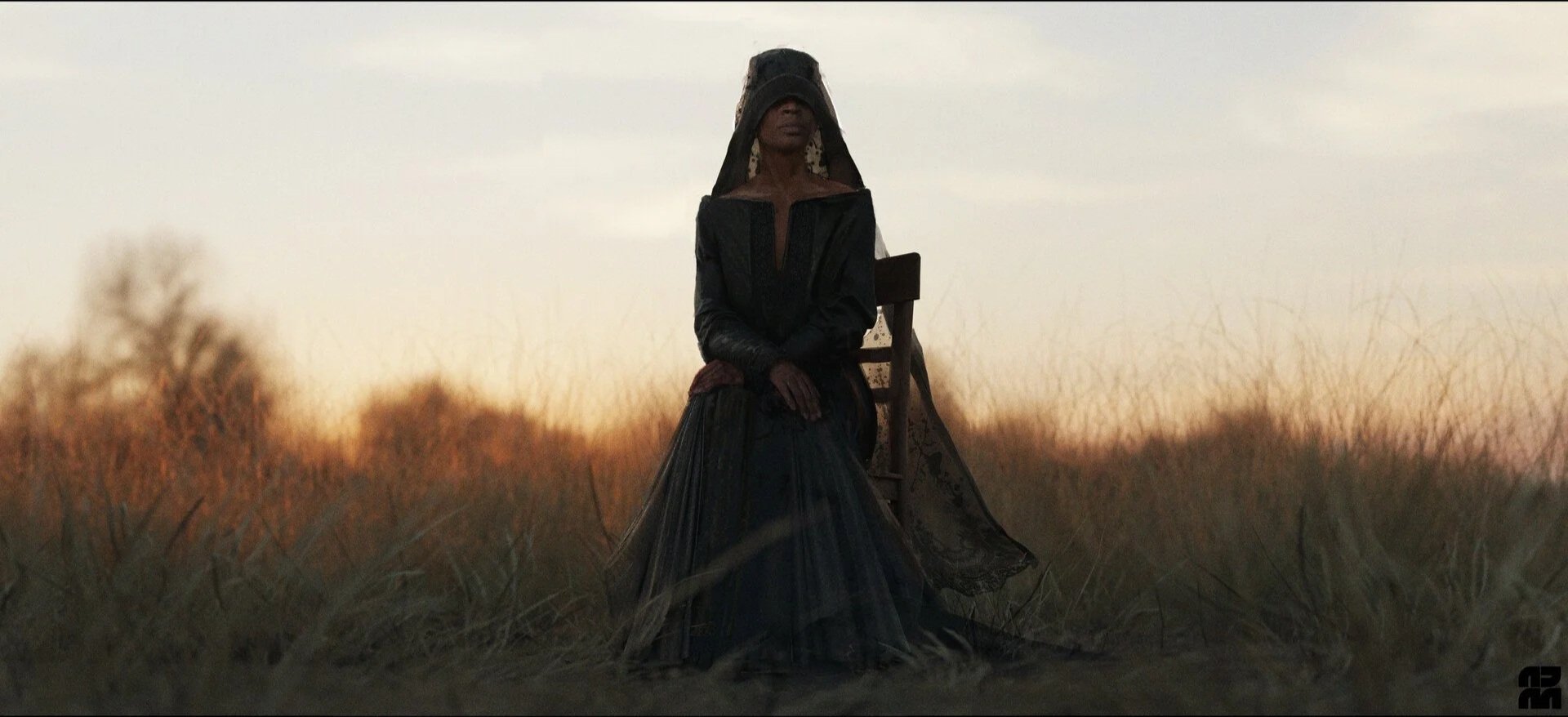 A woman in dark, vintage-style clothing sits on a wooden chair in a field at sunset, wearing a large black hooded cape and lace earrings.