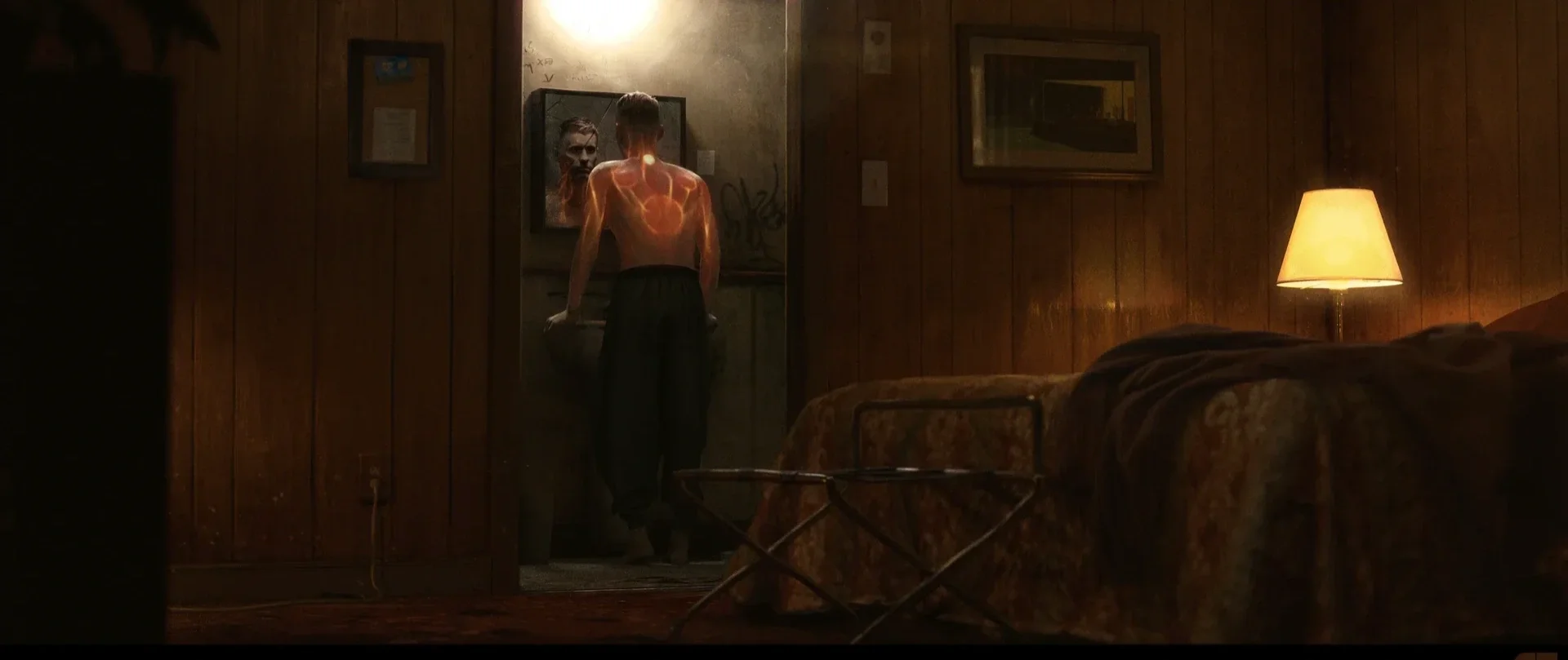 A man shirtless with glowing tattoos looking into a mirror in a dimly lit room with wood-paneled walls, a bed with a brown blanket, and a lamp.