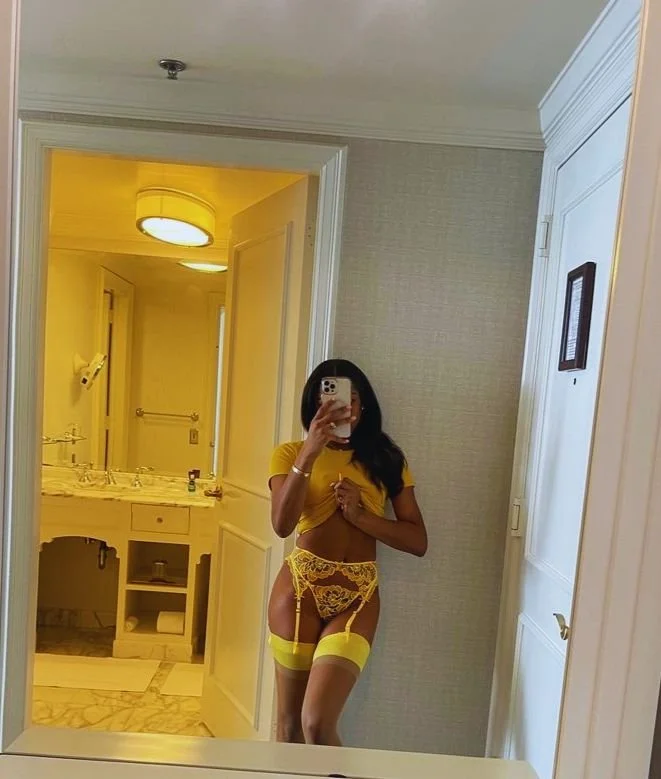 Woman taking a mirror selfie in a hotel room wearing yellow lingerie and thigh-high stockings