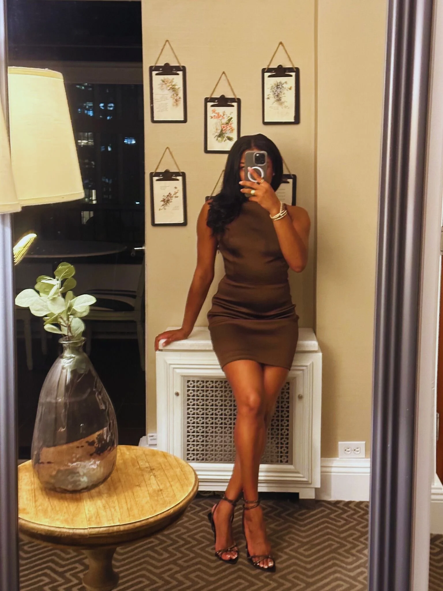A woman in a brown dress and high heels takes a mirror selfie indoors, standing with one hand on a white decorative radiator cover and the other holding a phone. The background has framed botanical prints on the wall, a window with a city view, and a