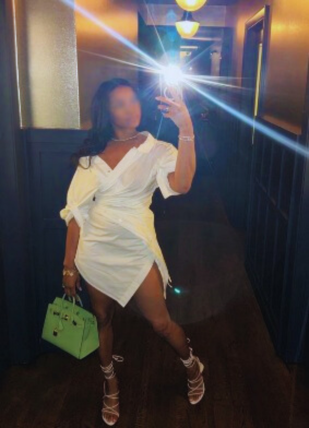 Woman taking a mirror selfie in a hallway, wearing a white dress and high heels, holding a green handbag.