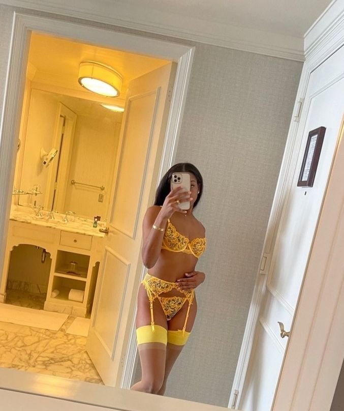 Woman in yellow lace lingerie and thigh-high stockings taking a mirror selfie in a hotel room.