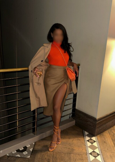 A woman standing indoors next to a railing, holding a glass of wine in one hand and a clutch purse in the other. She is wearing an orange top, a beige trench coat draped over her shoulders, a high-slit skirt, and strappy high heels, with a small oran