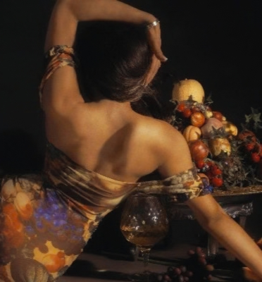A woman with dark hair and jewelry poses with her back to the camera, reaching over a table decorated with a variety of fruits and a glass of wine, in a dimly lit setting.
