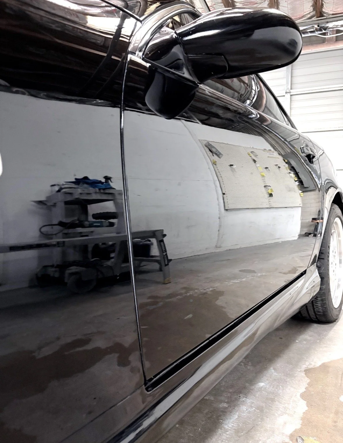 A close-up of a black car's shiny exterior side reflecting the workshop interior, including a workbench and tools, with a garage door in the background.