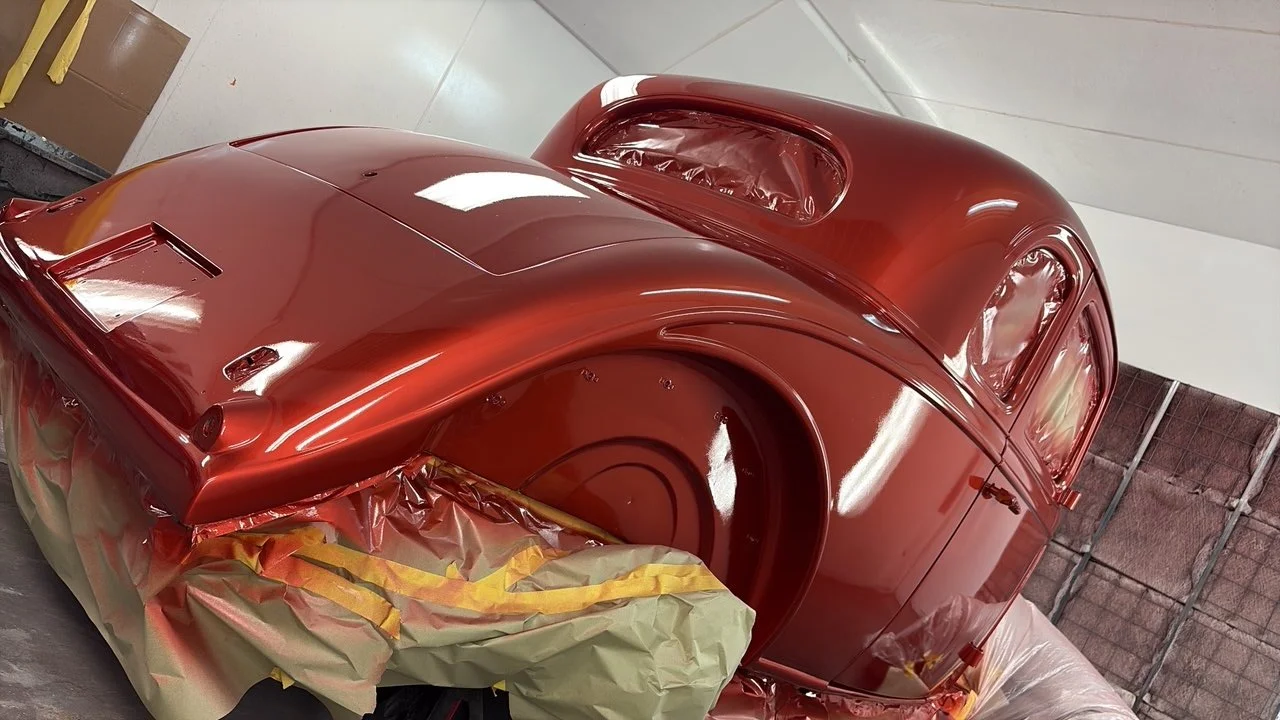 A classic car is being painted in a workshop. The car is coated in a shiny red paint, with masking paper and tape around certain areas for painting.