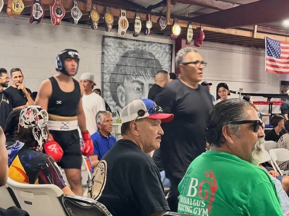 A boxing gym with several people seated and standing, some wearing boxing gloves and headgear. There is a large mural of a young boy's face on the wall, boxing championship belts hanging from the ceiling, and an American flag in the background.