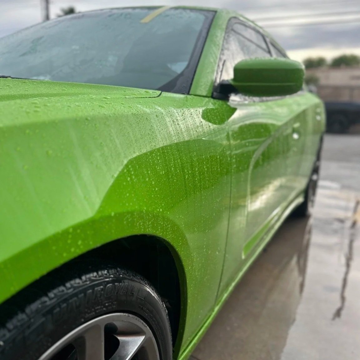 Close-up of a bright green sports car with water droplets on its surface, parked outdoors on a wet surface with a cloudy sky in the background.