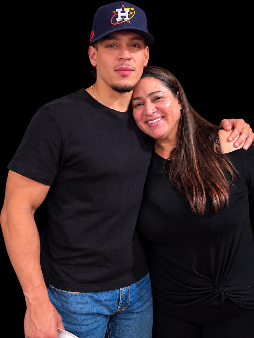 A young man wearing a Houston Astros baseball cap and a black t-shirt stands next to a woman with long brown hair, smiling, with her arm around him, against a black background.