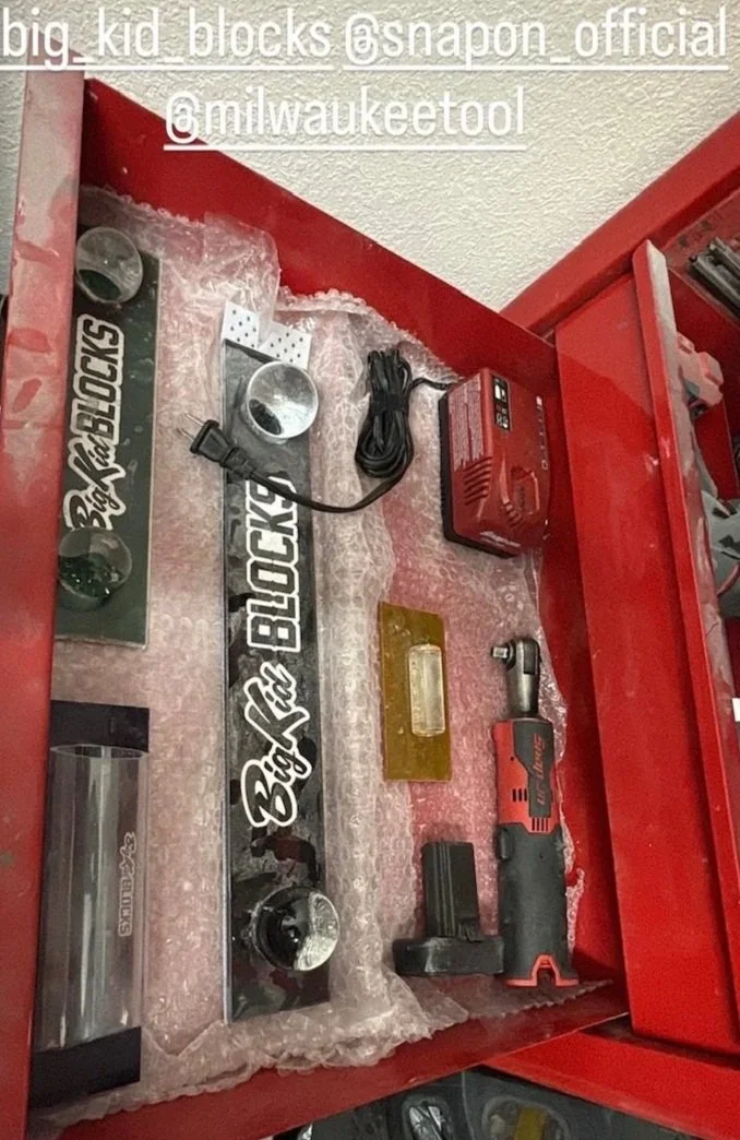 A red tool chest drawer containing a cordless drill, a metal sign reading 'Big Kid Blocks', a red power supply, a black cord, a small metal cylinder, and a brass-colored object with a transparent window.