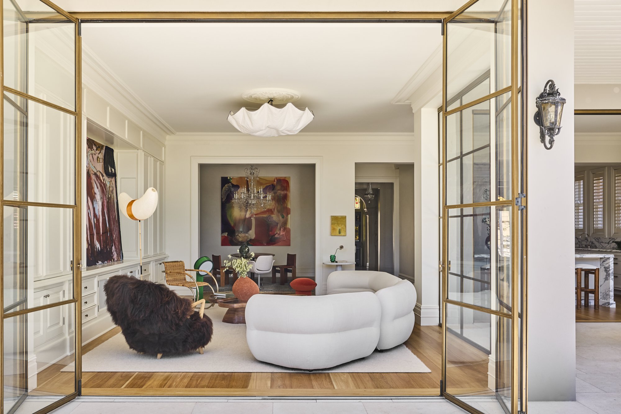 Living room with white curved sofa, furry brown armchair, wooden coffee table, rattan chairs, and artwork. Glass doors with gold frames open to the room.