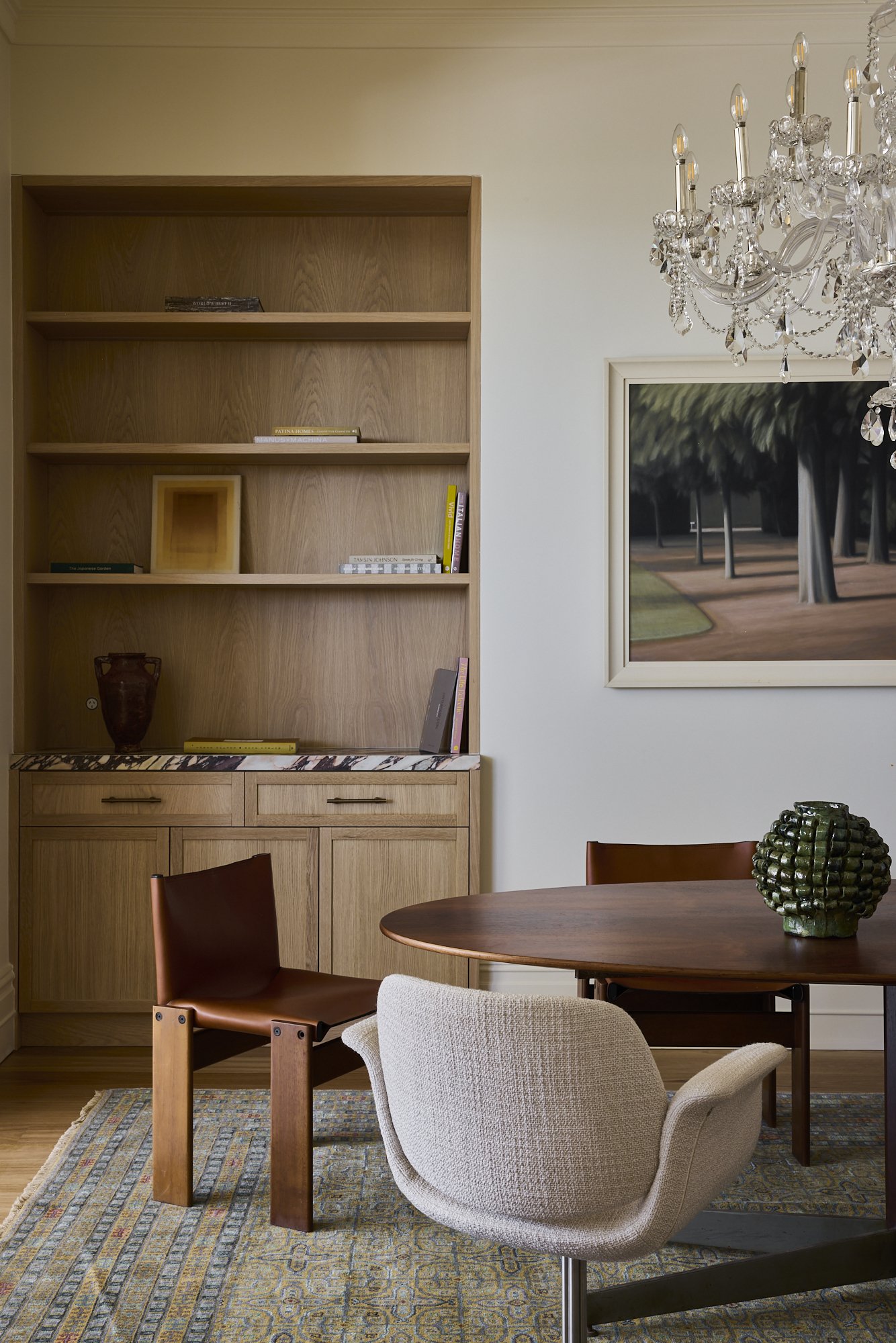 A room with a wooden bookshelf, a round wooden table with a green ceramic vase, and chairs including a beige upholstered chair and a brown leather chair. There is a large framed painting of trees on the wall and a crystal chandelier hanging from the ceiling.