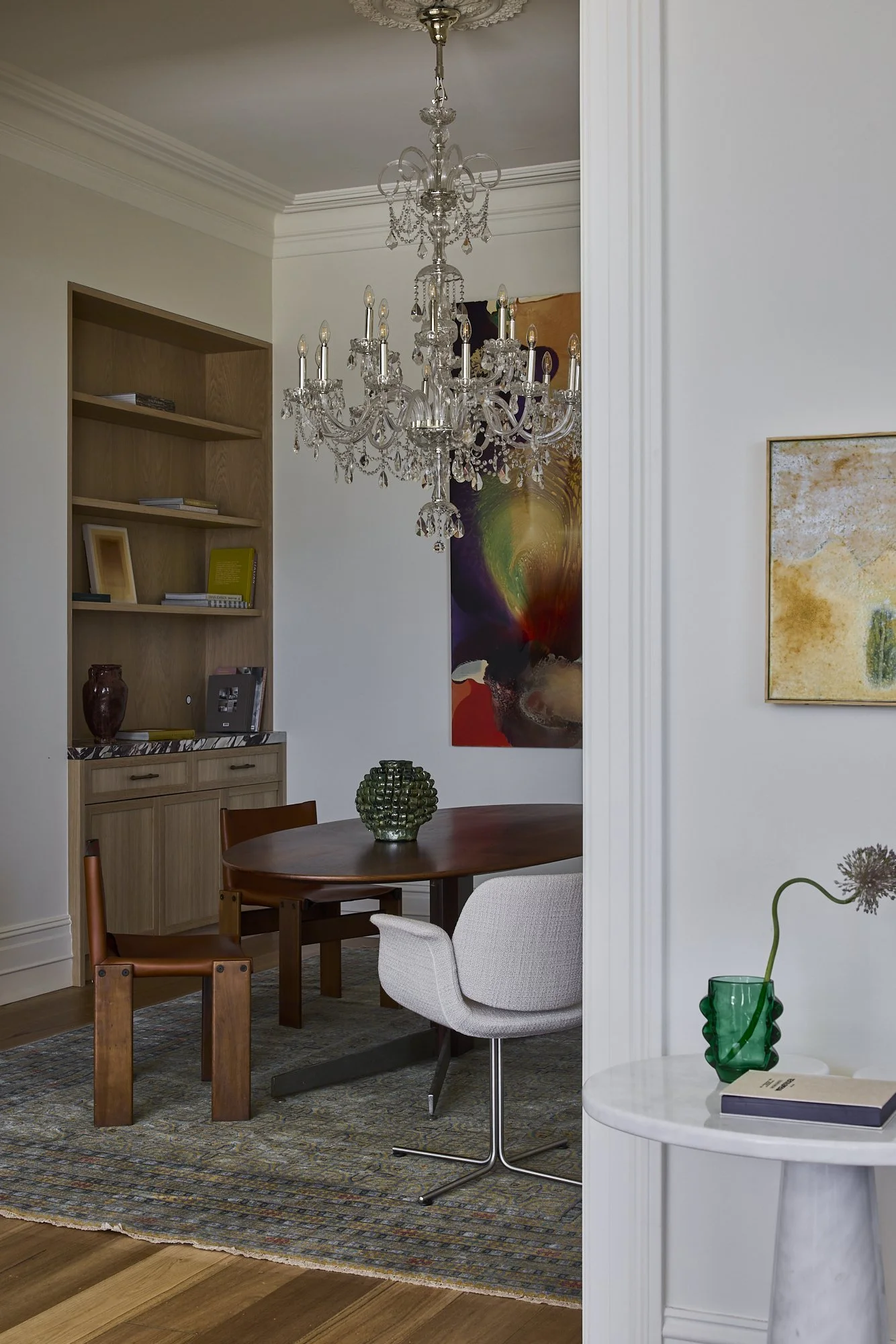 A dining room with a crystal chandelier, a wooden oval table, and a mix of chairs including an upholstered white chair, a brown wooden chair, and a small side table with a green vase and a book.