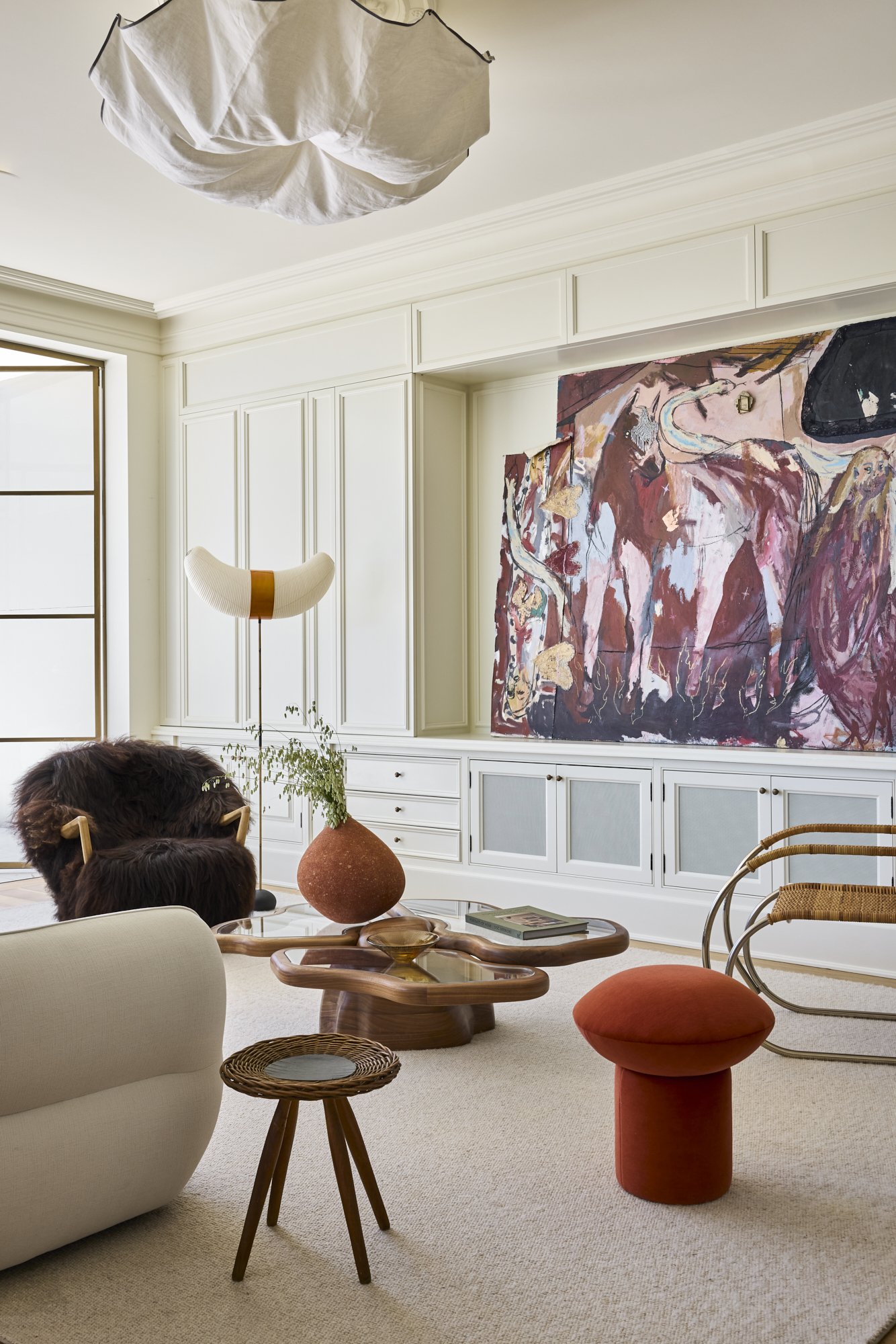 Living room with white built-in cabinetry, a large abstract painting, a cream sofa, a wooden coffee table, a textured terracotta vase with greenery, a modern sconce, a rocking chair with rattan details, and a plush orange ottoman.