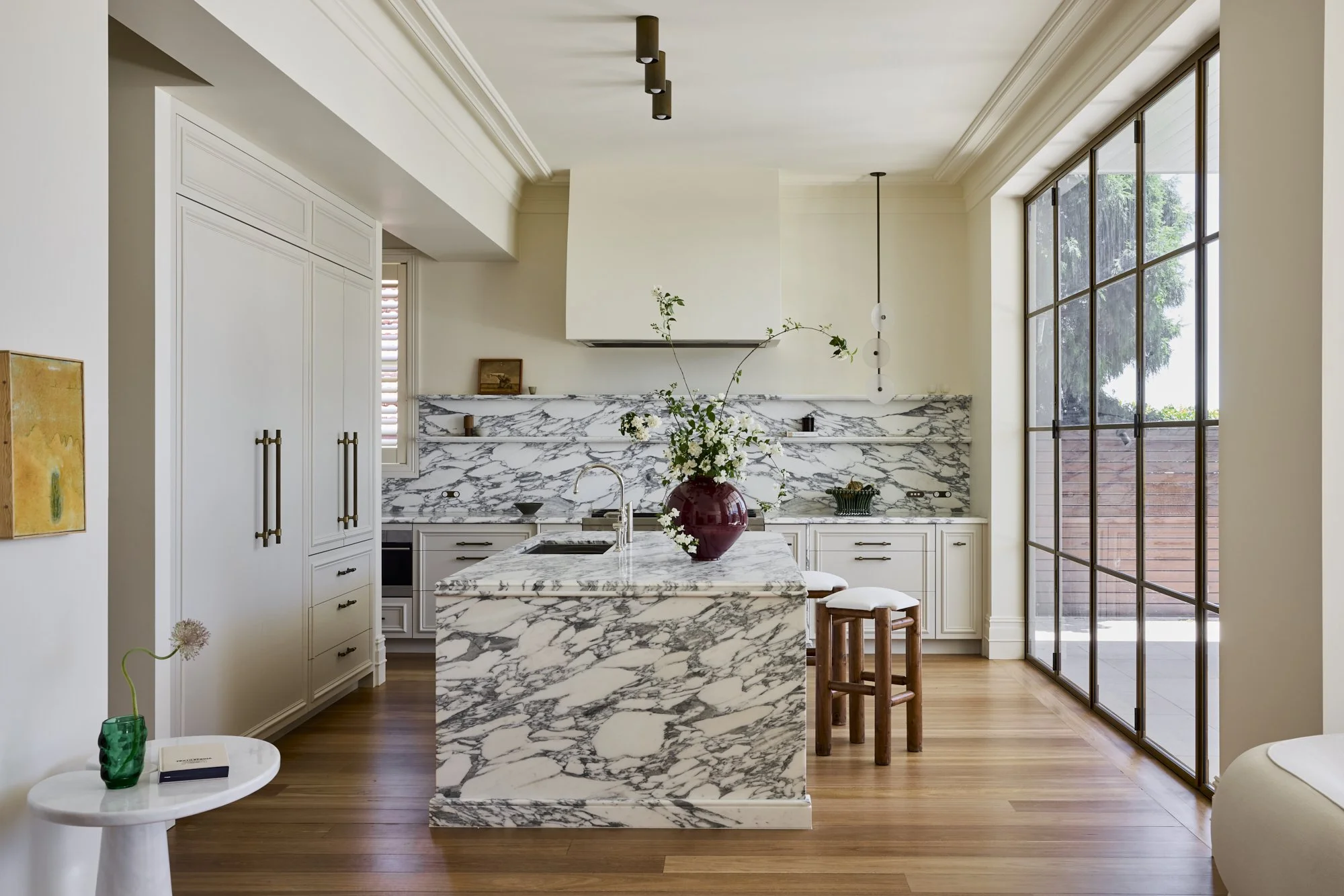 Modern kitchen with white cabinets, marble countertops, and a marble kitchen island. There is a large window on the right, and a vase with flowers on a small white table to the left.