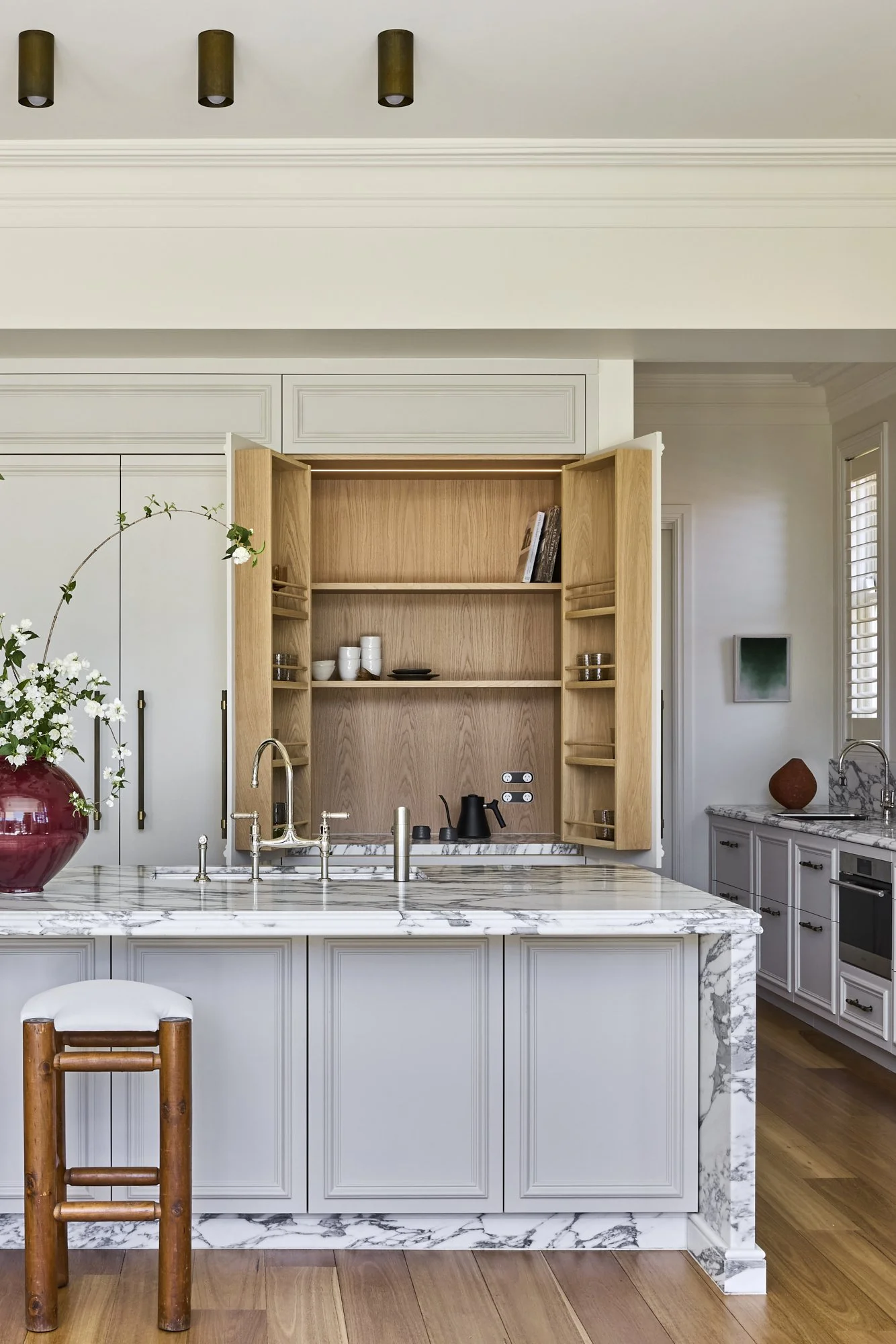 A spacious kitchen with white cabinets, a marble island with a built-in sink, a wooden open cabinet, and hardwood floors. There is a potted plant with white flowers in a red vase, and black kitchen appliances on the marble backsplash.