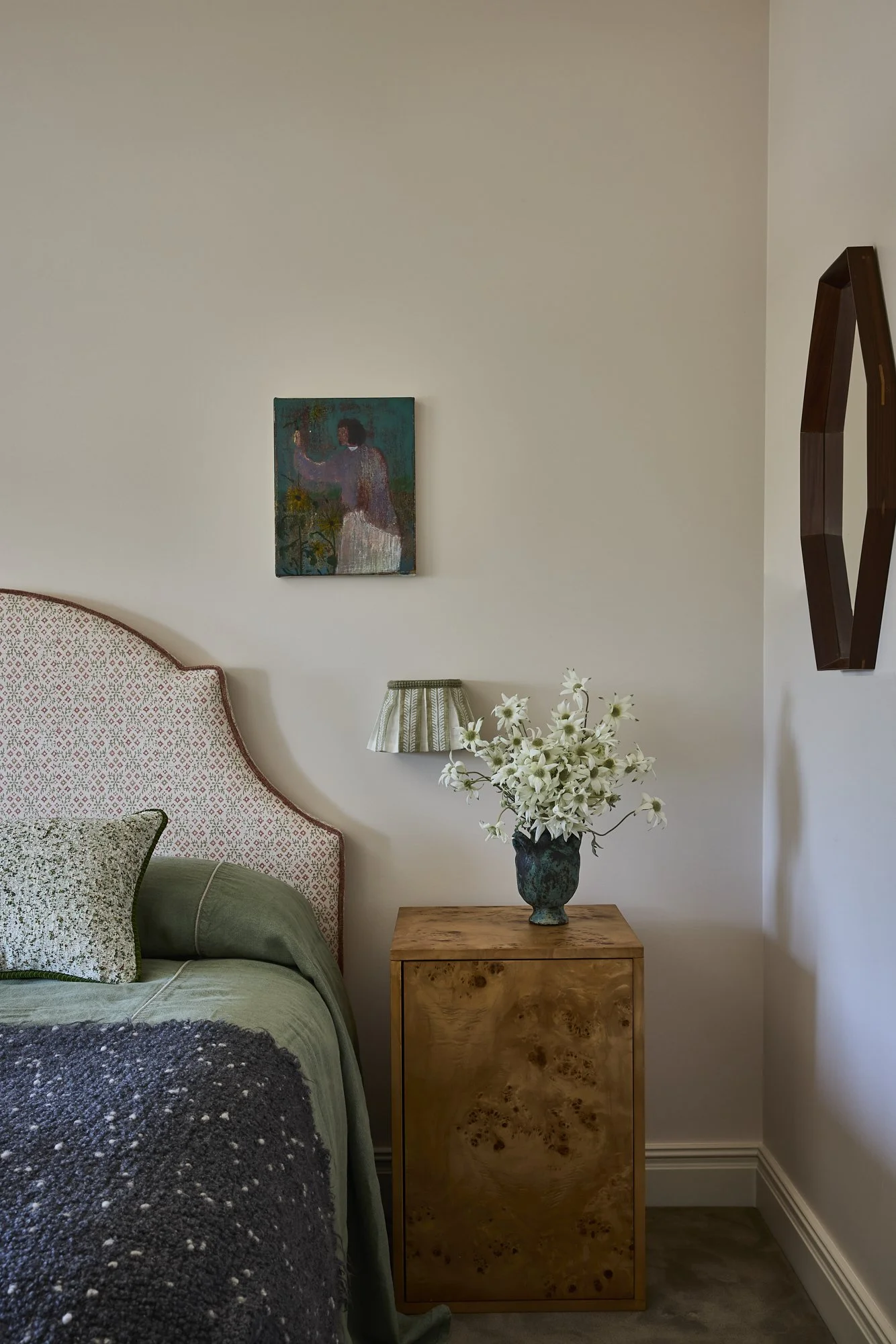 A bedroom corner with a bed, green bedding, a patterned pillow, and a wooden nightstand. The nightstand has a vase with white flowers. Above is a small wall lamp and a picture of a person with back turned, holding something, on the wall.