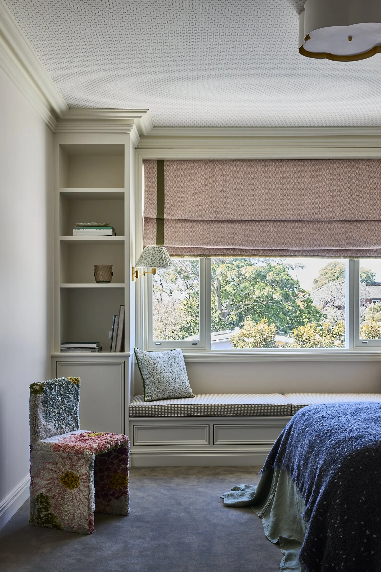 A cozy bedroom with a large window featuring a pink window shade, a built-in window seat with pillows, a patterned armchair, a partial view of a bed with a dark blue blanket, and built-in shelves with books and decor.