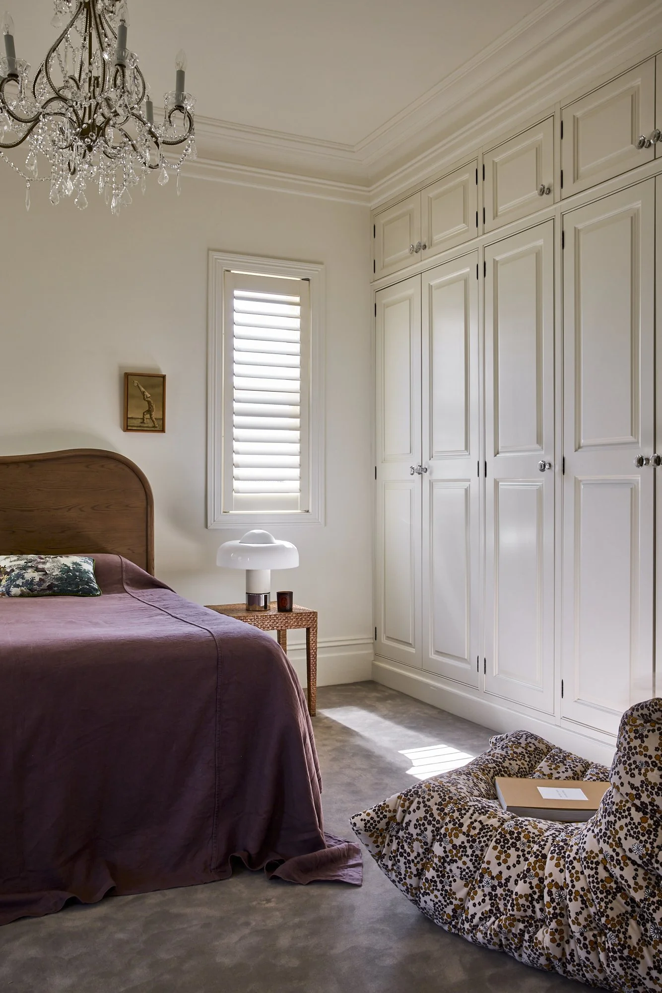 A bedroom with a chandelier, built-in white wardrobe, bed with purple bedspread and pillow, small bedside table with a white lamp and candle, window with white blinds, and a leopard print bean bag on the carpeted floor with a book on it.