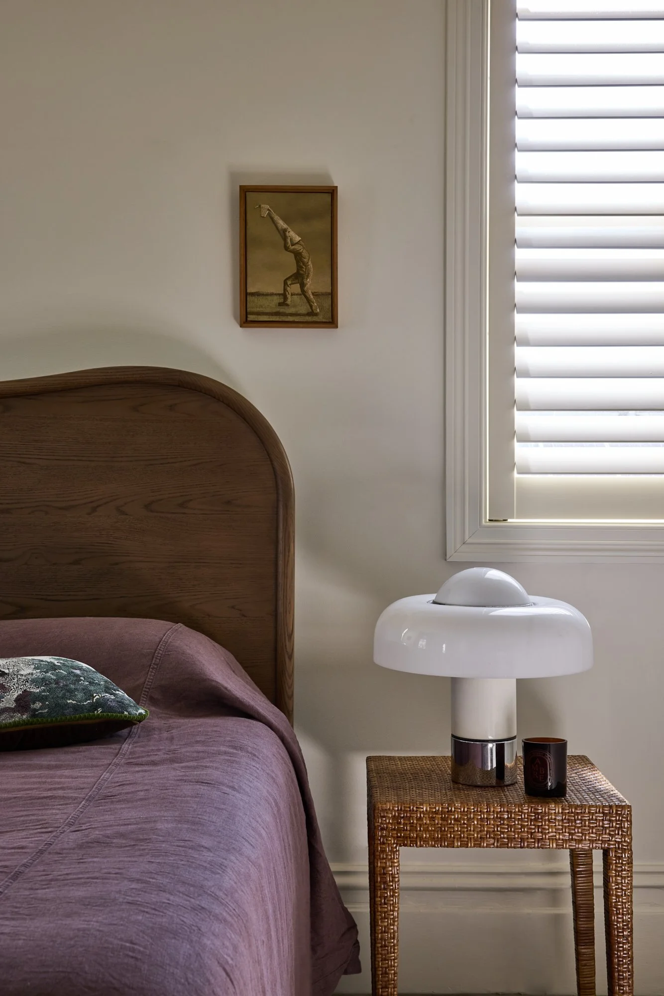 A bedroom with a wooden bed frame and mauve bedding, a bedside table with a white mushroom-shaped lamp, a black mug, a window with white blinds, and a framed picture of a person performing a handstand on the wall.