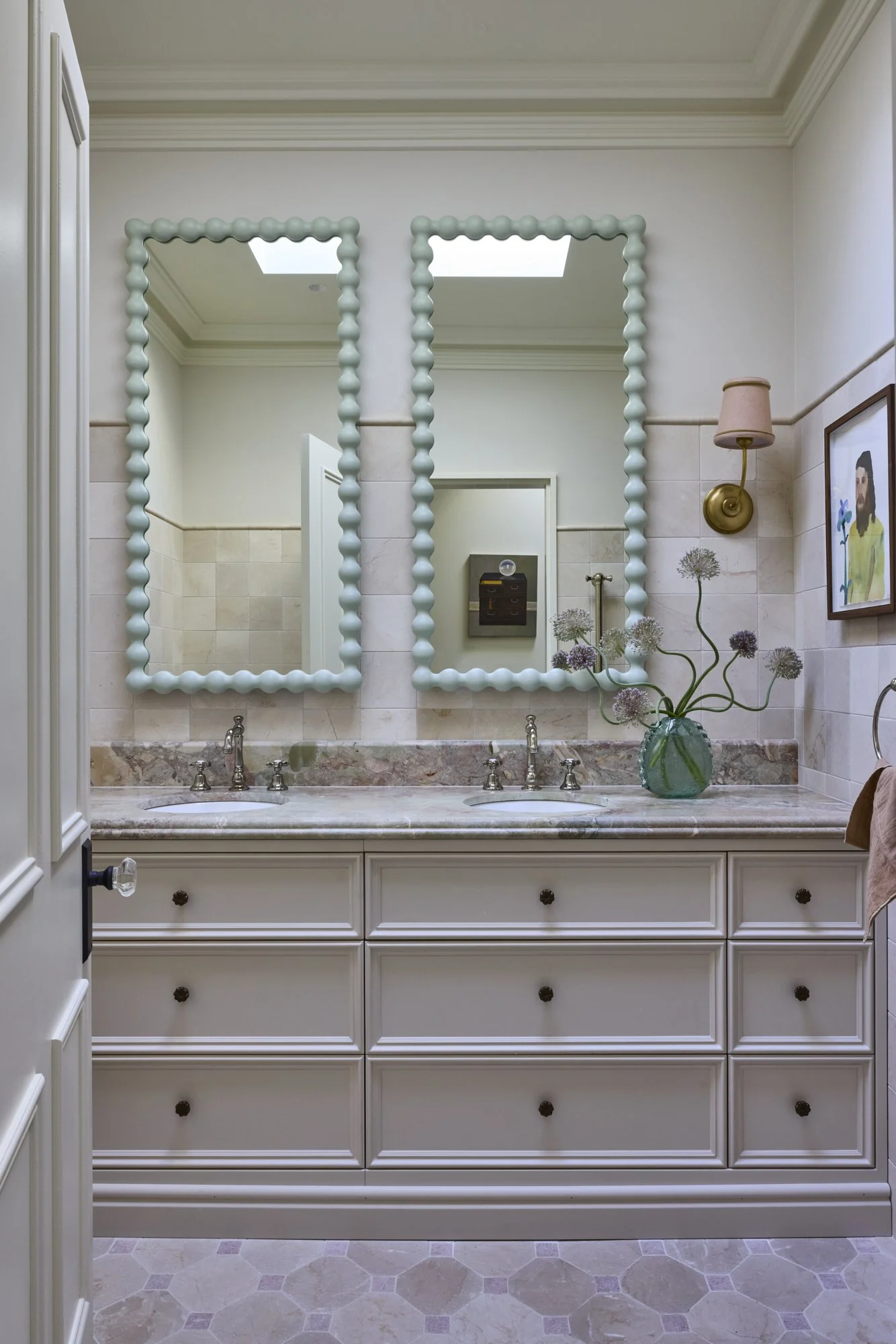 A bathroom with two decorated mirrors above a double sink vanity, a vase of flowers, gold wall light, and framed artwork.