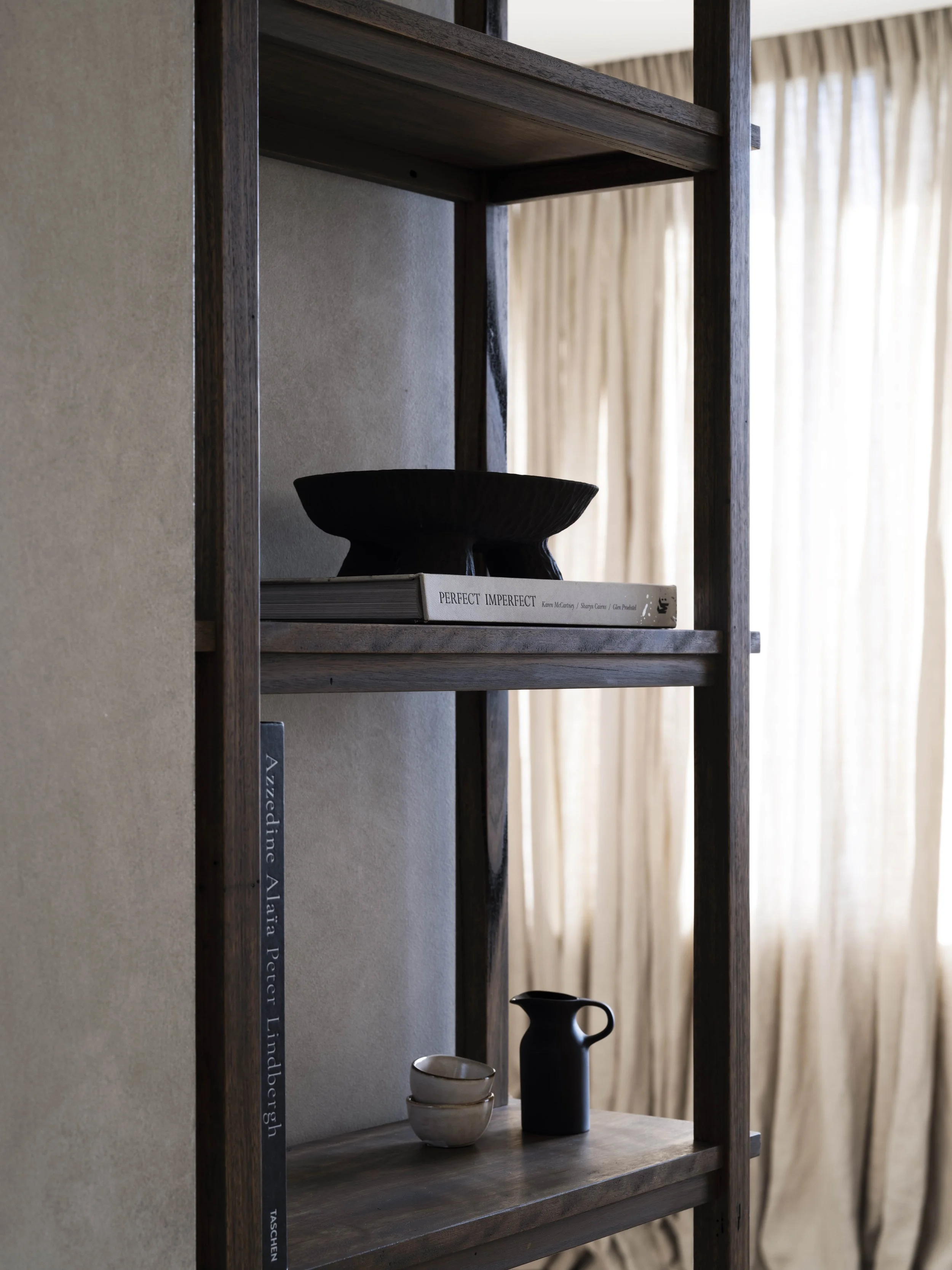 A wooden shelf containing a black bowl on top of a book titled 'Perfect Imperfect,' two small white bowls, and a black pitcher, with beige curtains in the background.