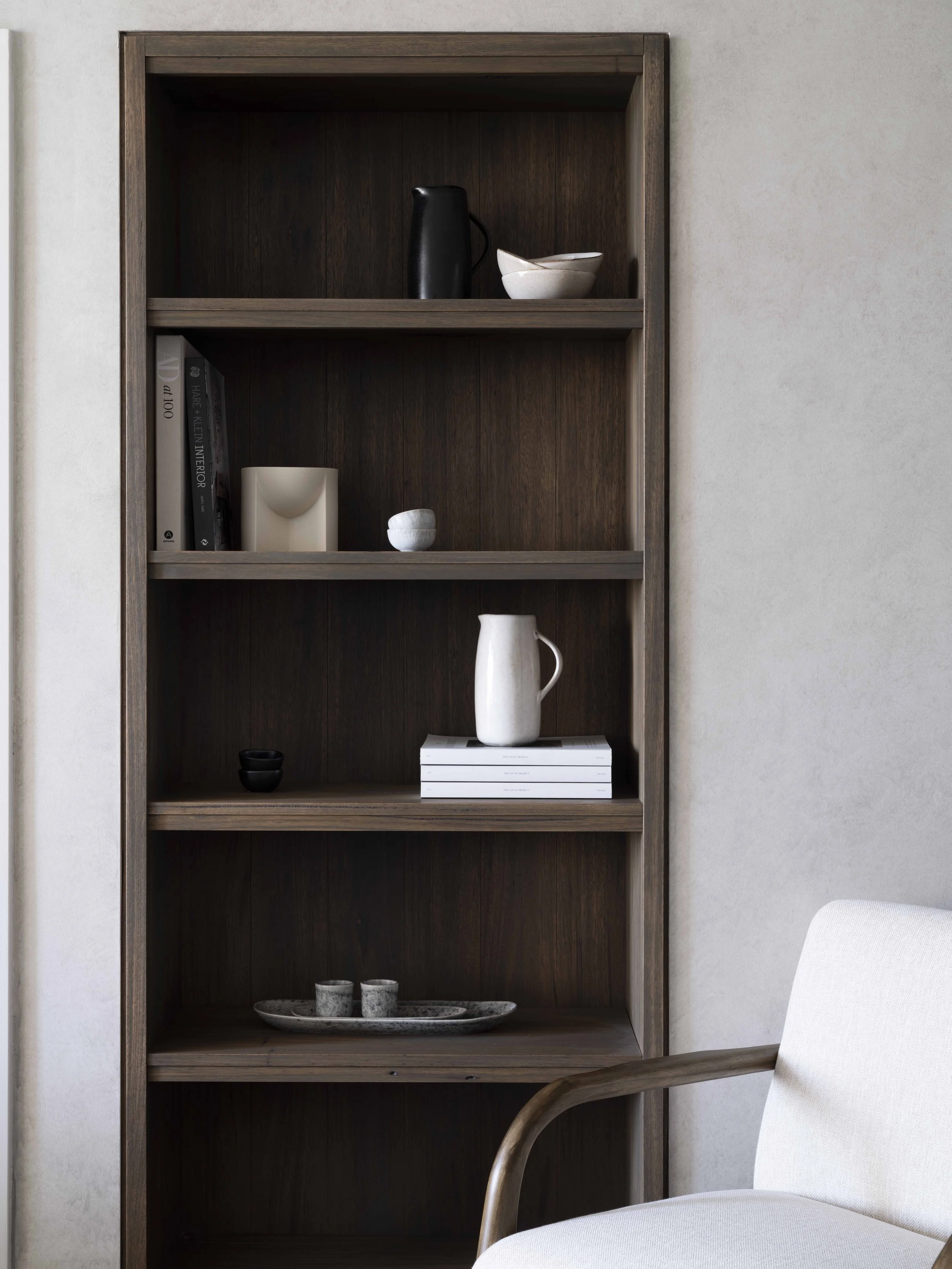 A dark wood bookshelf with five shelves filled with decorative objects, books, and dishes, next to a beige armchair.