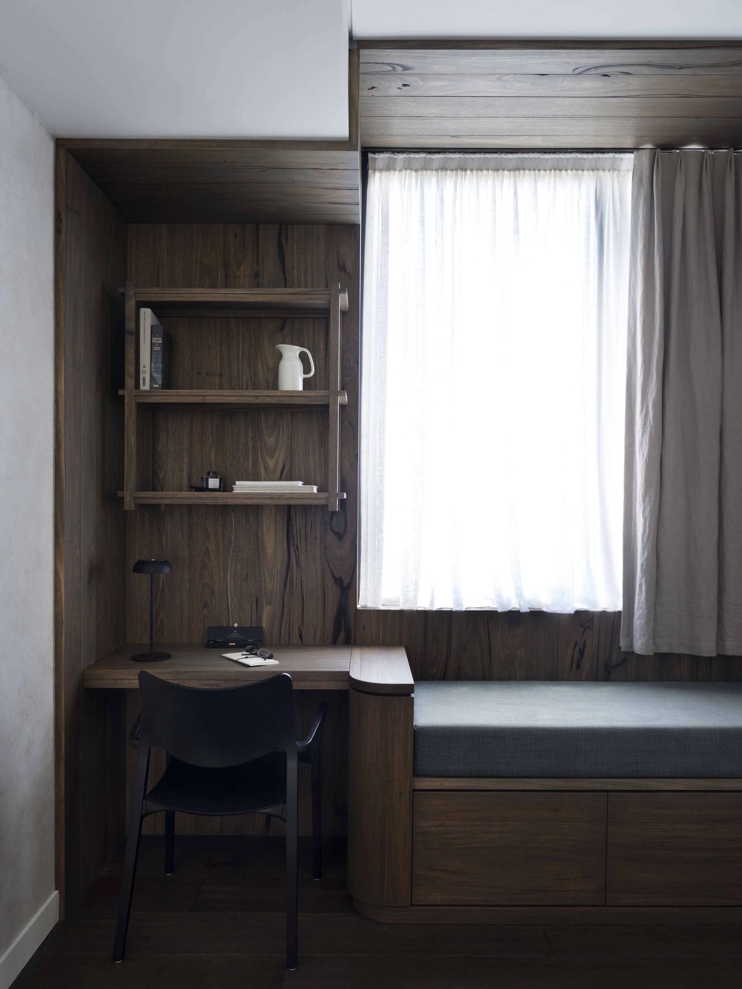 A corner of a cozy room with a wooden desk, a black chair, a bookshelf with decorative items, and a window with white curtains.