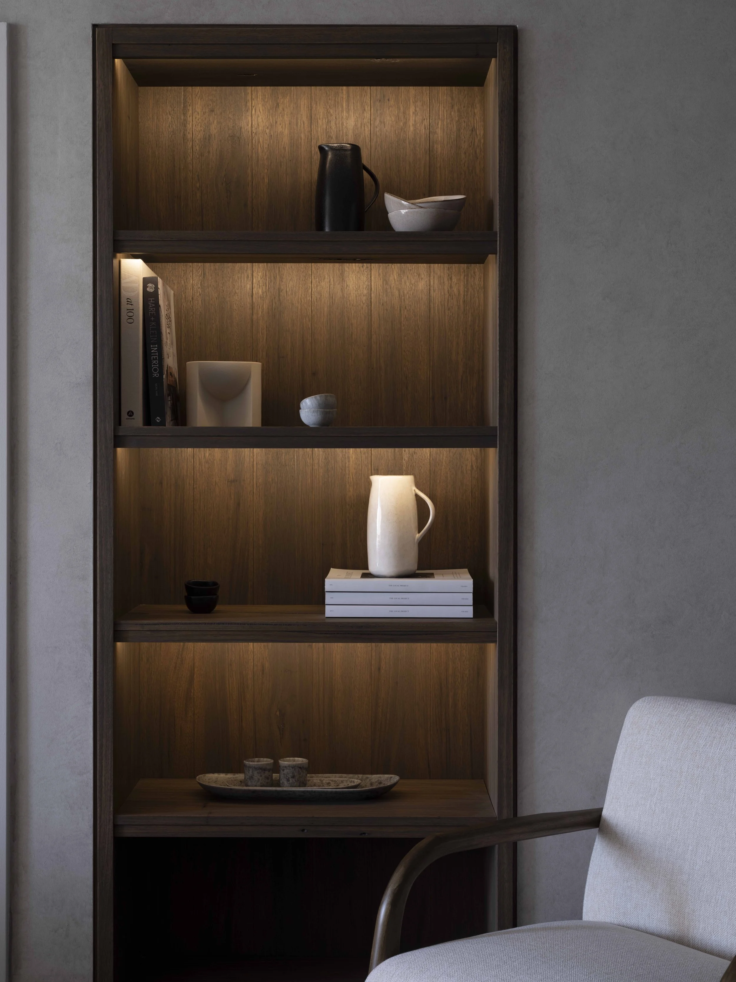 A tall wooden bookshelf built into a wall, containing decorative vases, bowls, books, and small objects, with integrated lighting, next to an upholstered chair.