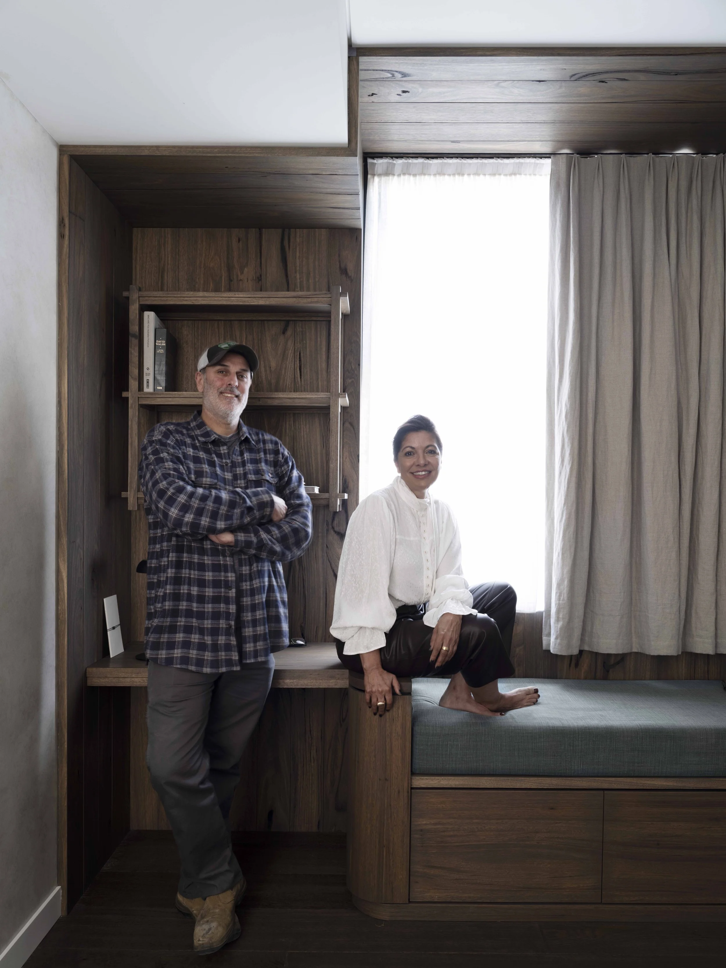 A man and a woman in a room with wooden walls and a large window with curtains. The man is standing with arms crossed, wearing a plaid shirt, gray pants, and a cap. The woman is sitting on a wooden ledge, wearing a white blouse, dark pants, and barefoot, smiling at the camera.