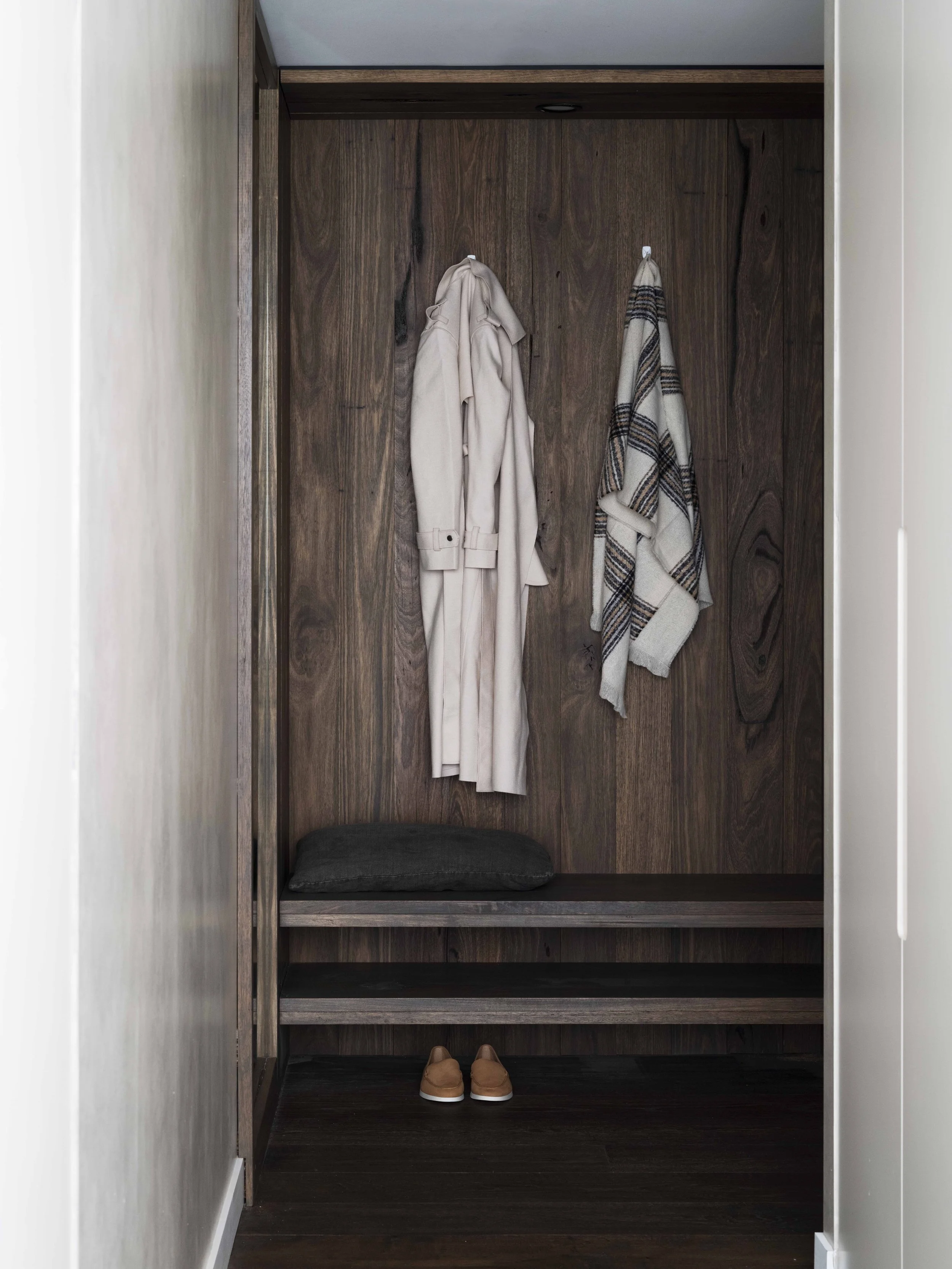 Empty wooden closet with a beige trench coat and plaid scarf hanging on hooks, a black cushion on a bench, and a pair of tan shoes on the floor.